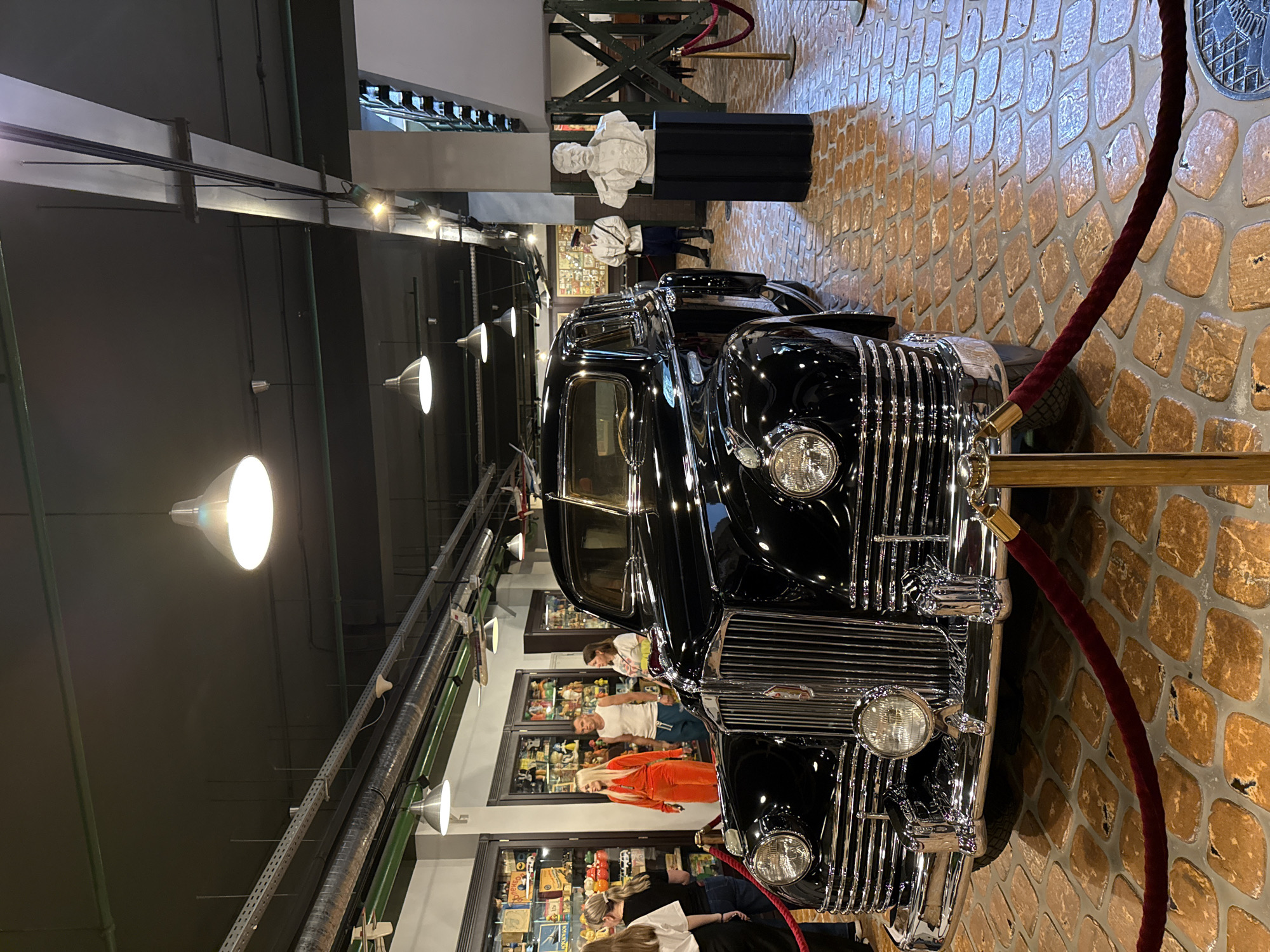 In the city of Arkhangel'skoe, Russia, within what appears to be a museum or exhibition space, a polished black vintage automobile is prominently displayed. The car, a large sedan with chrome accents on its grille, headlights, and bumper, is positioned on a floor paved with irregularly shaped, tan-colored stones. A deep red velvet rope, held by golden stanchions, cordons off the vehicle.
Behind the car, several individuals are visible. To the left, a group of people, including women wearing casual attire, are looking towards display cases filled with various items. Further back, a man in a white tank top and blue shorts stands next to a woman in an orange tracksuit. To the right of the car, a bust of a man is mounted on a black pedestal, and a figure dressed in a historical military-style uniform, possibly a guard or mannequin, stands behind it. The environment is illuminated by overhead lights, and the ceiling features exposed ductwork and lighting fixtures. Several model airplanes are suspended from the ceiling in the background.