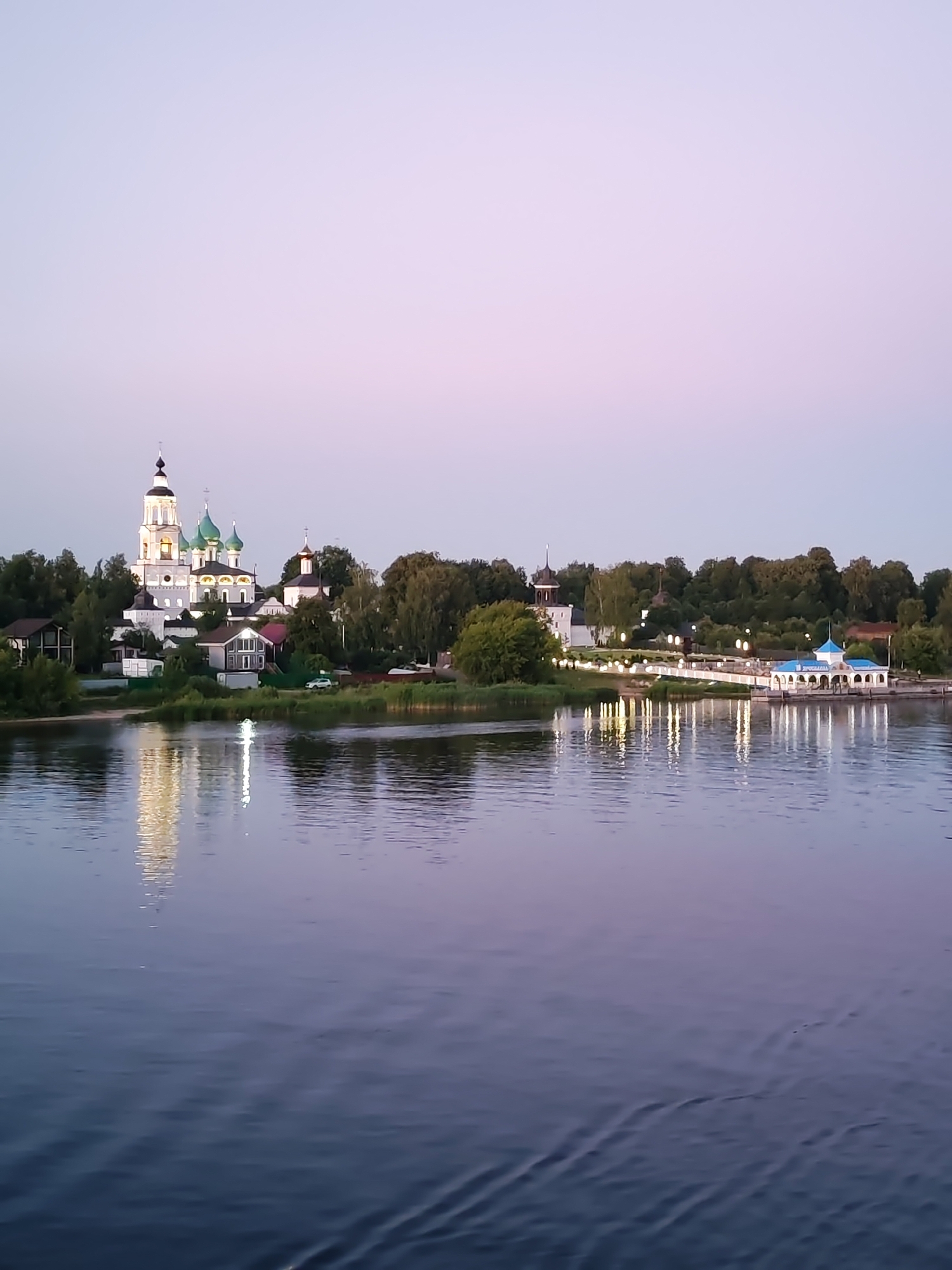 A wide shot captures a serene river scene at dusk. The water, dark blue with subtle ripples, reflects the twilight sky and the illuminated buildings on the opposite bank. A prominent white church with green domes stands tall amidst other structures and lush green trees. To the right, a covered walkway with a blue roof and arched openings is also visible, with lights reflecting in the water. The sky transitions from a pale purple at the horizon to a soft pinkish hue higher up. The overall atmosphere is peaceful and tranquil. The specific location is unknown.