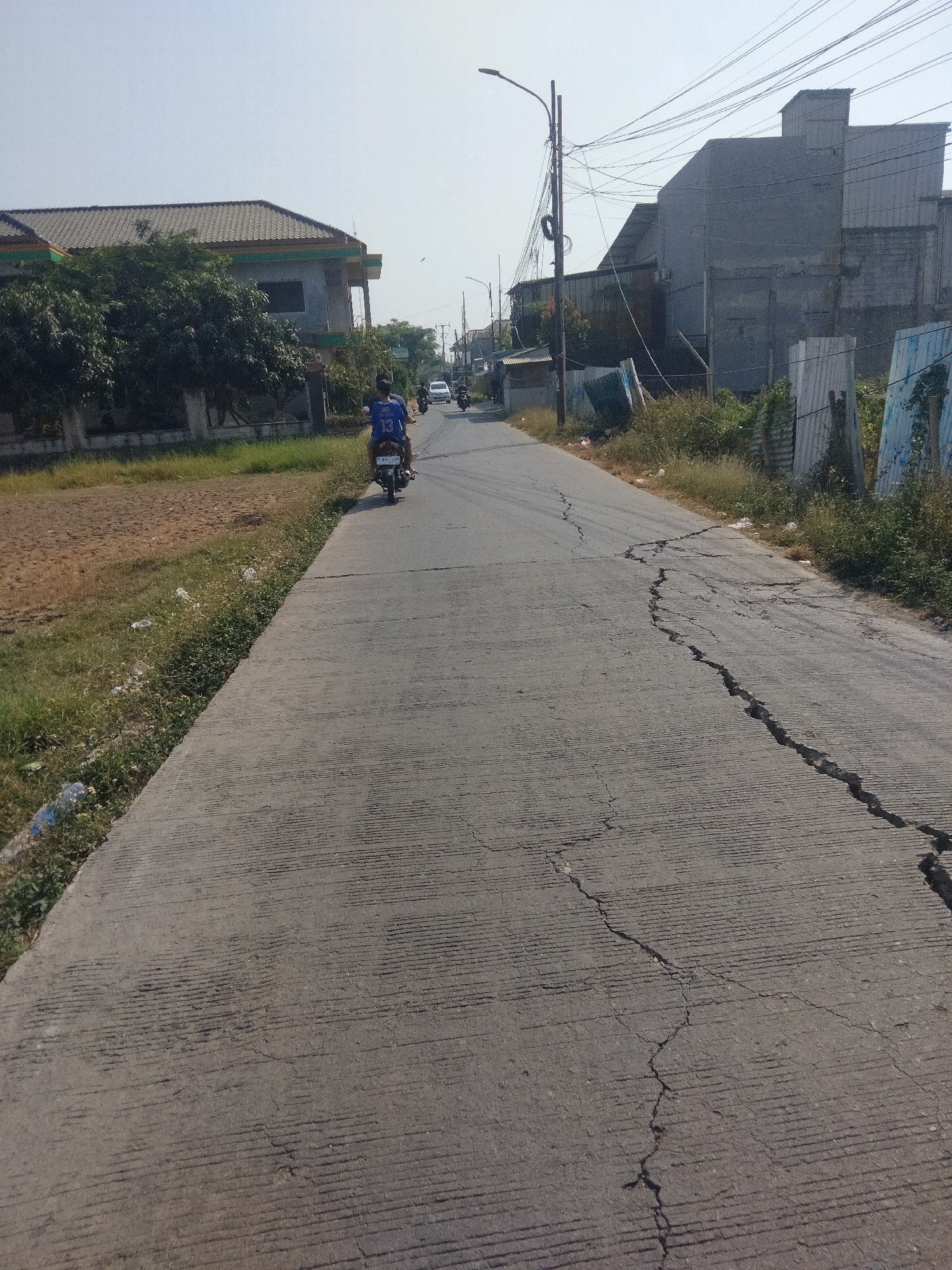 This image depicts a street scene in Suka Sari, Indonesia, on a clear, sunny day. The foreground shows a cracked concrete road with parallel grooves, indicating wear and tear. To the left of the road, there is a dirt field with sparse dry grass and some scattered plastic litter. A concrete sidewalk runs alongside the dirt field. Further down the road, a person is riding a motorcycle, wearing a blue shirt with the number "13" on the back. Buildings line the right side of the road, including a two-story structure with a gray facade and a tiled roof, and other industrial-looking buildings. Overhead, a network of electrical wires crisscrosses the sky. The overall environment suggests an urban or semi-urban setting in Indonesia.