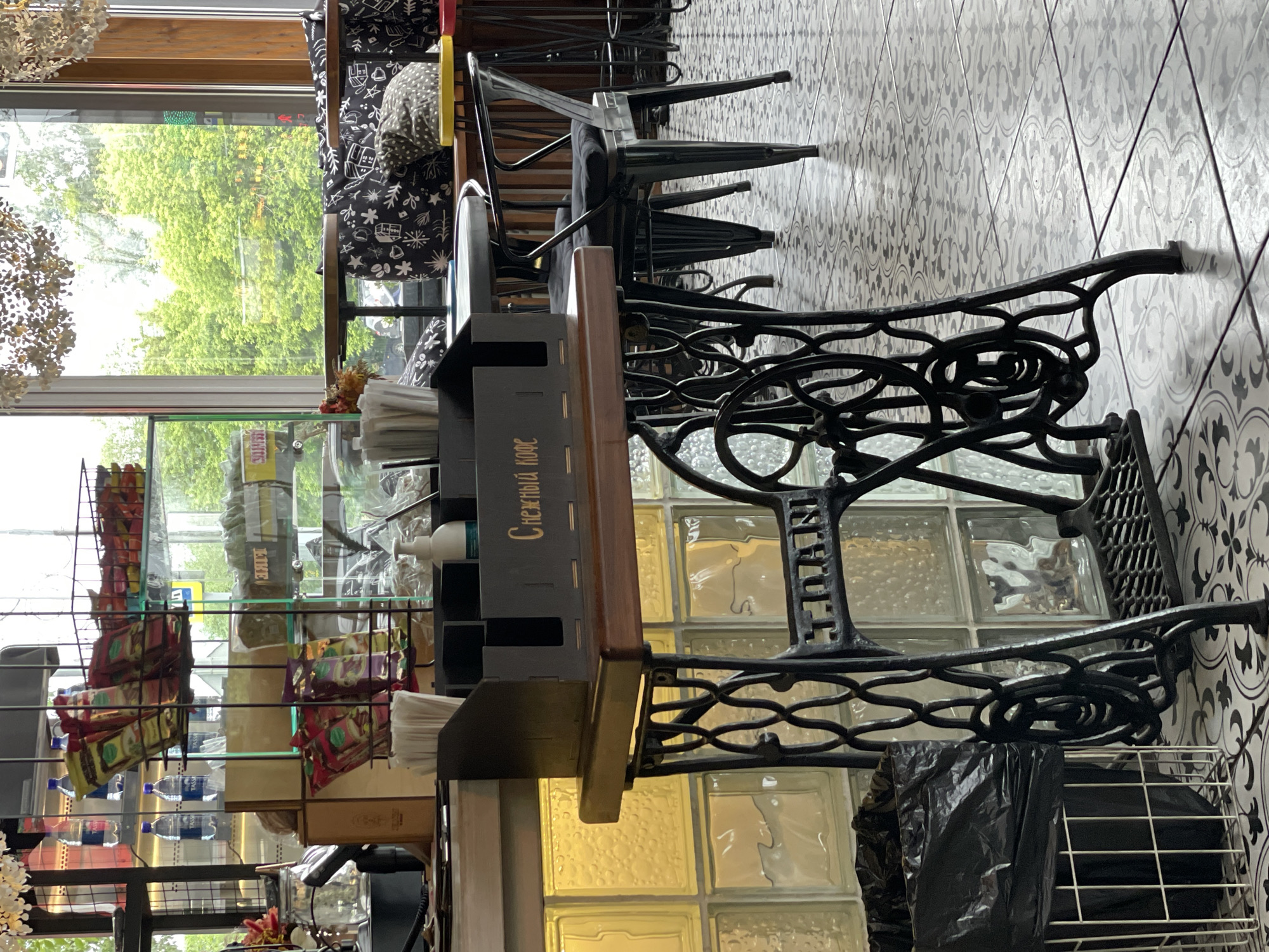 The image depicts the interior of a cafe, likely in Krasnoyarsk, Russia, as indicated by the Cyrillic text "Снежный кофе" (Snowy Coffee) on the dark wooden organizer. The scene is brightly lit, with natural light streaming in from a large window that offers a view of green trees, suggesting an outdoor setting.
In the foreground, a black metal sewing machine base serves as a unique table stand. It supports a dark wooden top with a wooden organizer containing napkins and other items. To the left of the sewing machine base is a white wire basket with a black trash bag.
Behind the table, several black chairs with dark cushions are arranged around small wooden tables, indicating seating areas for patrons. The floor is covered with a patterned tile in shades of gray and white.
In the background, shelves stocked with packaged goods, including bottles of water and bags of what appear to be snacks or coffee, are visible. A white pump bottle, possibly hand sanitizer or soap, is also present on a counter. The walls are partially clad in translucent, yellow-tinted glass blocks, adding to the ambient lighting. The overall impression is of a modern, slightly quirky cafe environment.