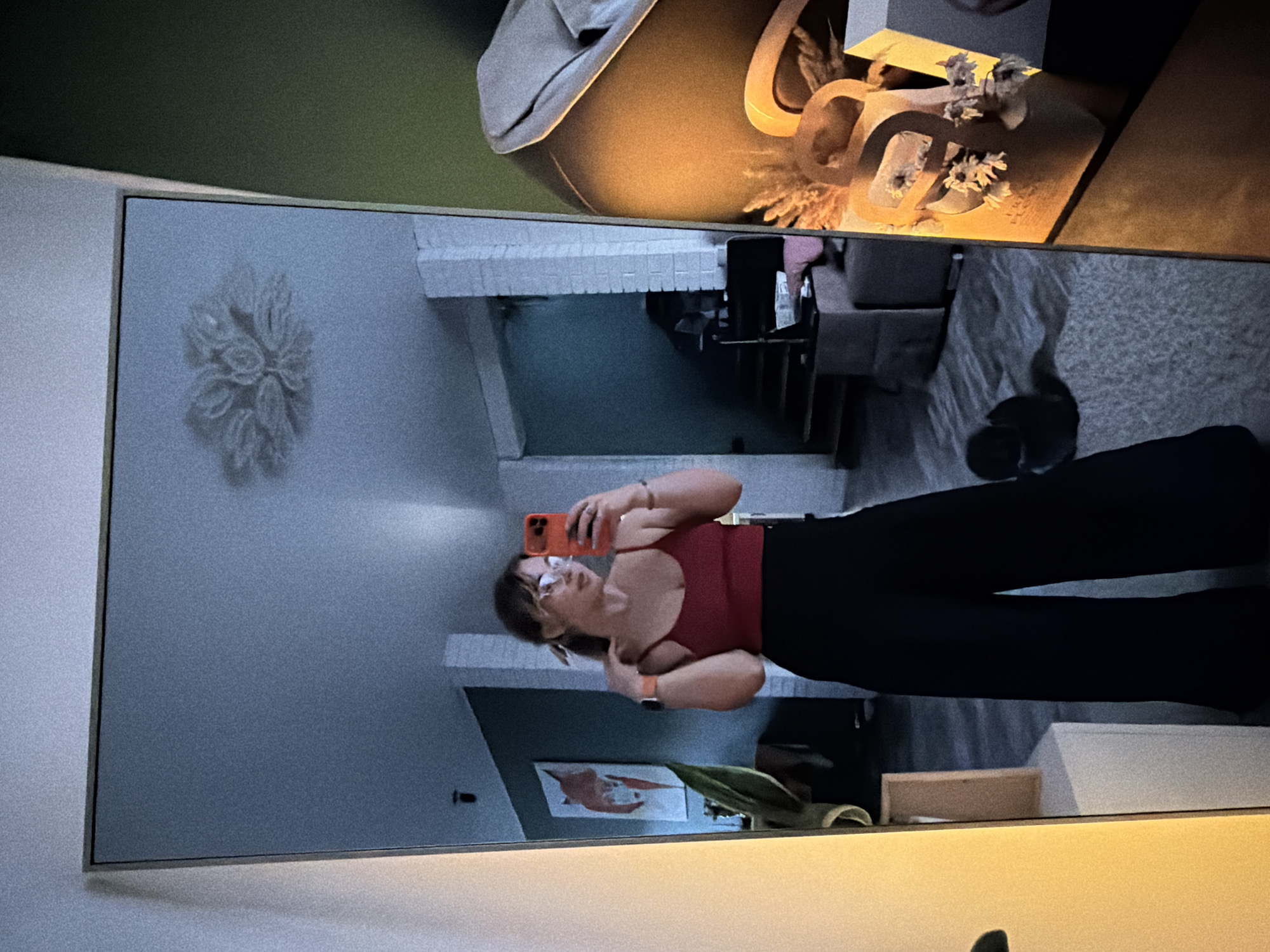 A medium shot shows a mirror reflecting a person taking a picture of themselves. The person, facing the mirror, wears a red tank top, black pants, and glasses, and holds an orange smartphone. The reflection shows a hallway with a white brick archway, a wall decoration of a white flower, and framed artwork on the lower right. There is a table and a lamp with decorations on top of it. The exact location within Большая Шильна, Russia, cannot be determined without further information.