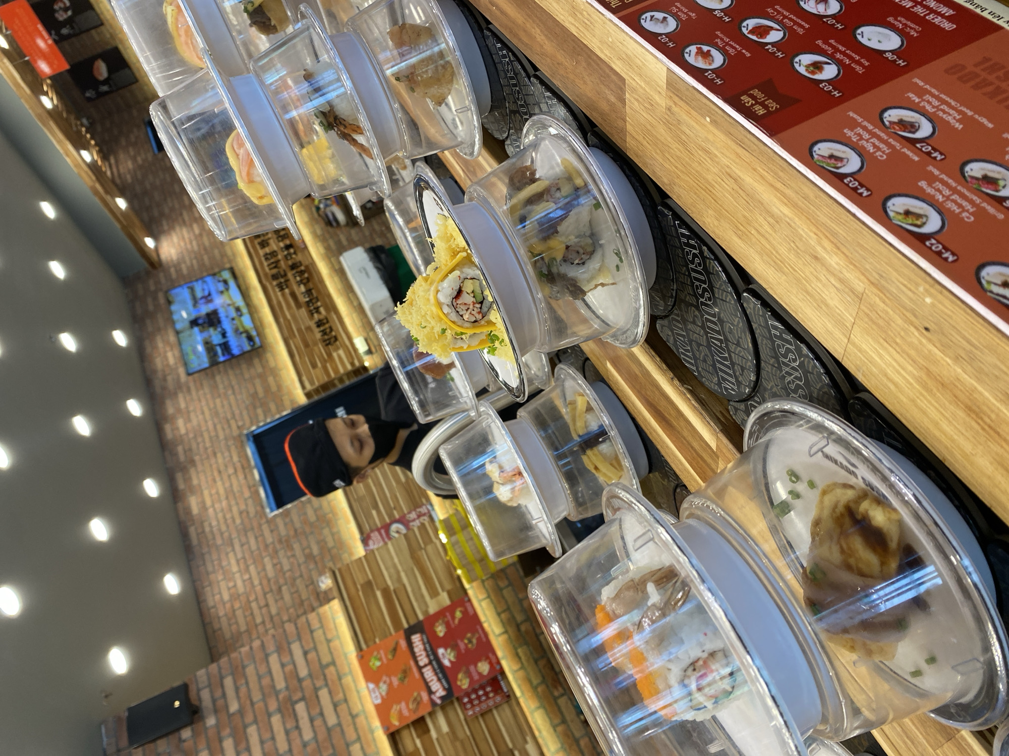 A conveyor belt sushi restaurant in Nha Trang, Vietnam, features a busy scene with various sushi dishes in transparent plastic containers moving along a wooden conveyor belt. A restaurant employee wearing a black cap and face mask stands behind the counter, seemingly preparing or serving food. The restaurant interior is decorated with brick walls and illuminated by overhead lights. On the wall, a digital screen displays images, possibly menus or advertisements. Menus with Vietnamese text are visible on the side, indicating specific sushi items and prices. The overall ambiance suggests a lively and efficient dining establishment.