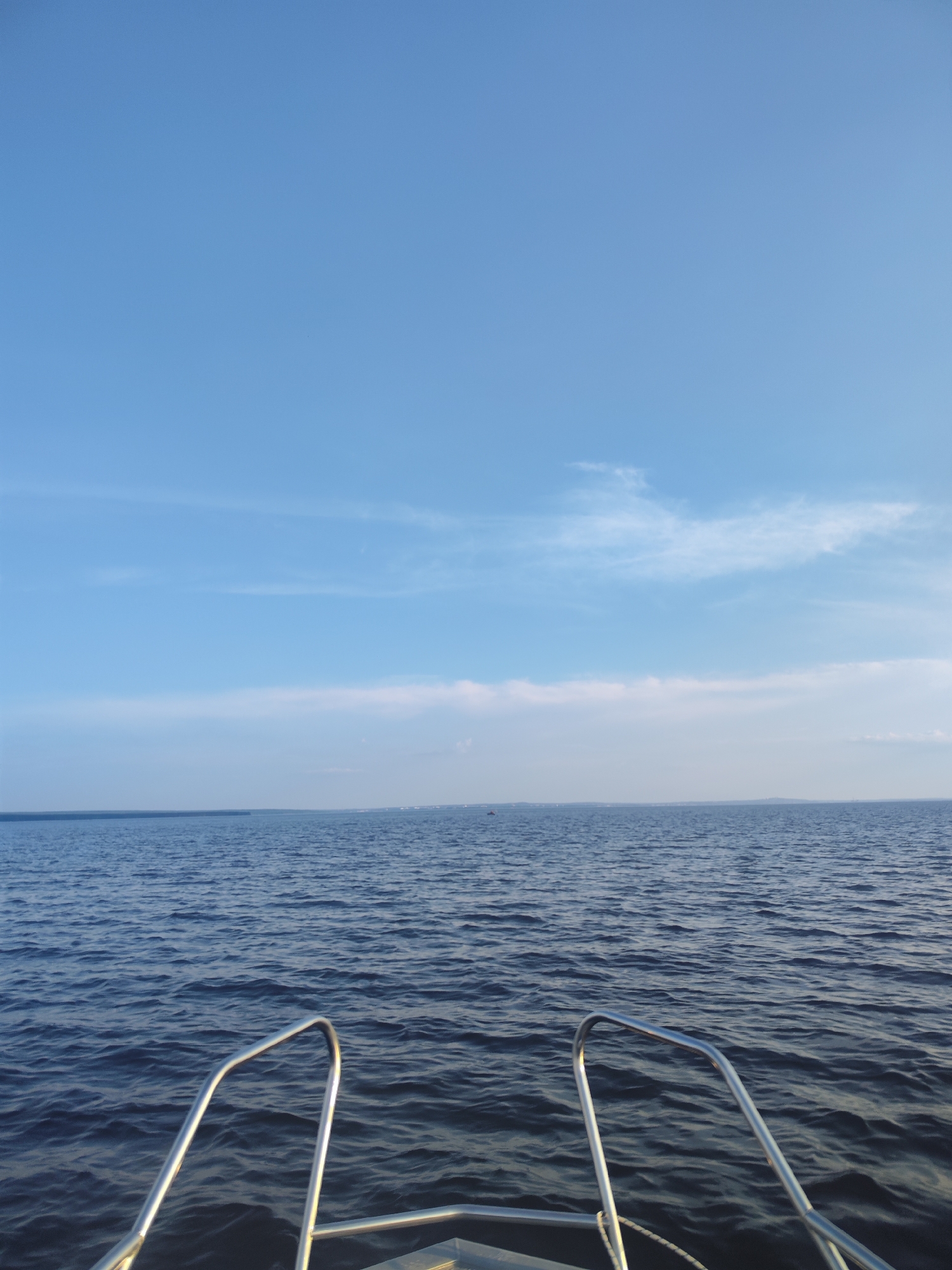 The image is a point-of-view shot from the front of a boat on a body of water. Two metal railings are visible in the foreground, extending from the boat's deck. The water is dark blue with gentle waves. In the distance, a faint line of land with some structures is visible on the horizon under a clear blue sky with scattered white clouds. The location is described as Galye, Russia. There are no people or animals visible in the frame.