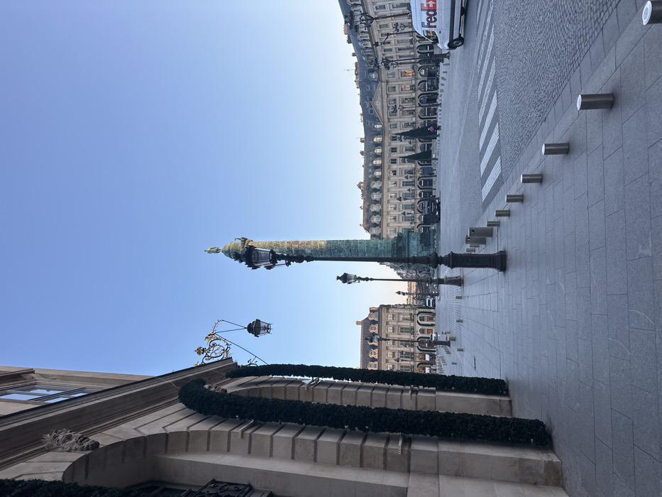 The image depicts an exterior view of Place Vendôme in Paris, France, oriented towards the central Vendôme Column under a clear blue sky.
In the left foreground, the facade of a classical building, constructed from light-colored stone, exhibits an arched entryway adorned with two vertical green garlands and a decorative mascaron carving. An elaborate wrought-iron street lamp bracket, featuring two lanterns, extends from this building. A grey tiled sidewalk runs along the building, transitioning into a grey cobblestone street marked by a white striped pedestrian crosswalk. A line of short, cylindrical, silver bollards demarcates the edge of the sidewalk and extends into the midground.
The bronze-clad Vendôme Column, topped with a statue, dominates the central midground. A tall, multi-lanterned black street lamp stands prominently in front of the column. The background is framed by the uniform, multi-story classical buildings characteristic of Place Vendôme.
On the right side of the street, a white FedEx delivery van with red and blue branding is visible. Several dark vehicles are sparsely distributed across the square. Two indistinct human figures are visible in the distant center of the square. The scene is illuminated by natural daylight, casting shadows and indicating clear weather. The square appears sparsely populated.