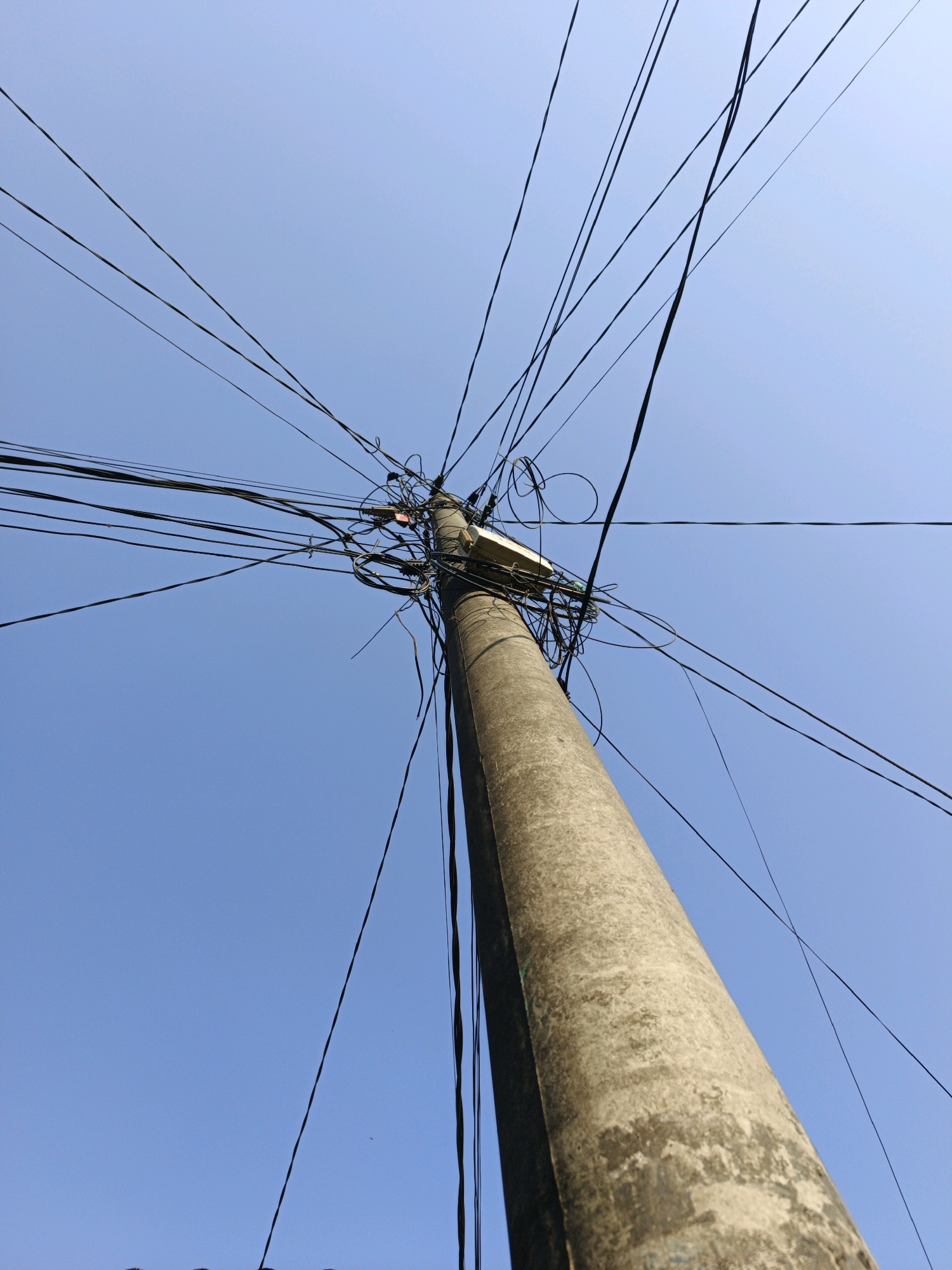 A low-angle, outdoor, full shot captures a utility pole against a clear blue sky in Ketegan, Indonesia. The concrete pole is heavily draped with a dense network of black electrical wires, some appearing tangled and haphazard. A white fixture, possibly a street light or transformer, is attached to the upper portion of the pole. The wires fan out in multiple directions across the frame. The sky is cloudless and bright, suggesting a daytime scene. No people are visible.
