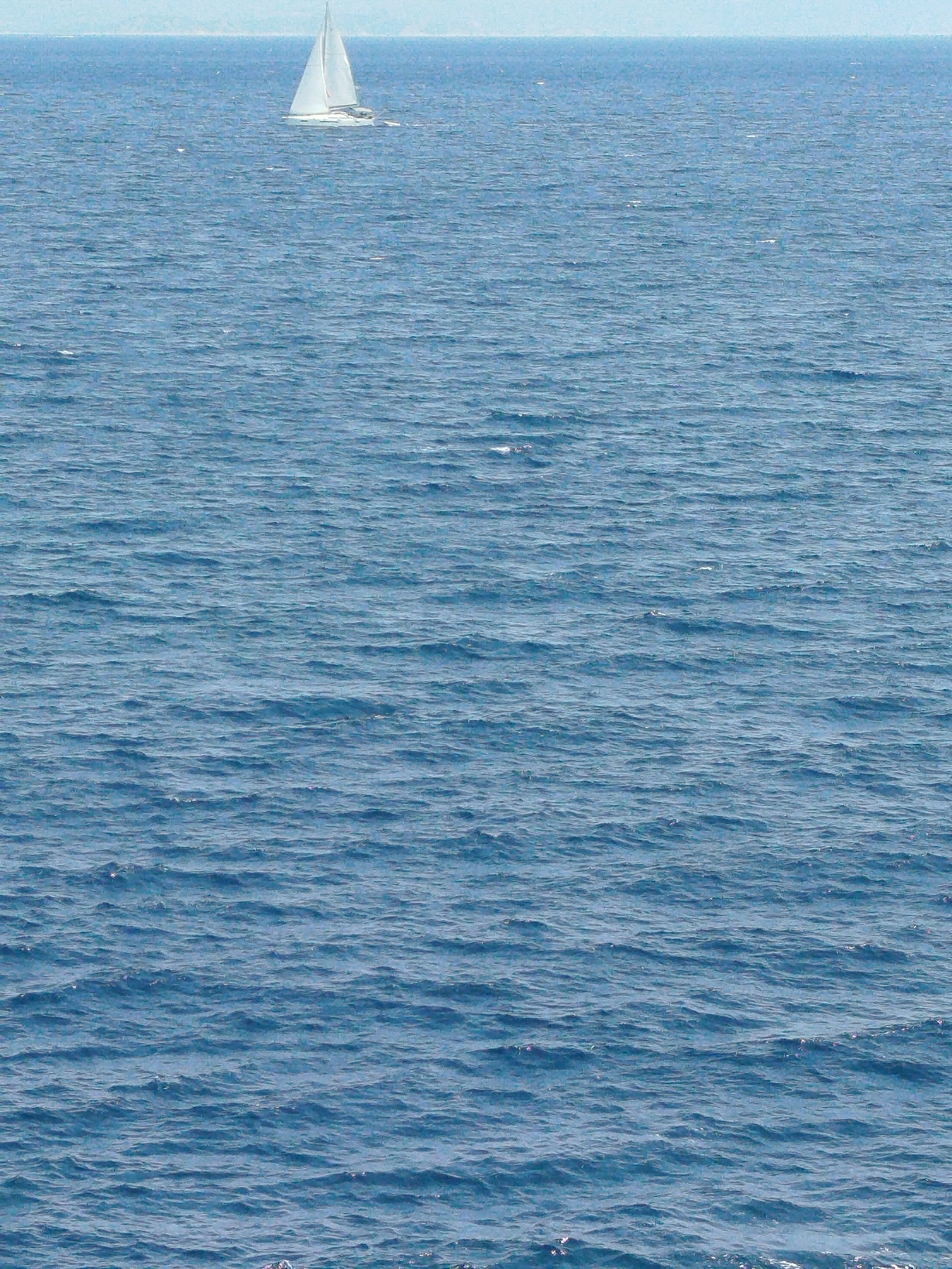 A white sailboat with its sails up is depicted on the blue sea, heading to the right. The boat is positioned in the upper left quadrant of the image. The sea is slightly choppy with small waves. The horizon line is visible in the distance, with a hazy, light-colored landmass beyond. The sky above is a pale blue. The overall scene is serene and captures a moment of sailing on a clear day. This scene is likely located within the waters of Sithonia Municipal Unit, Greece, a region known for its beautiful coastline and sailing opportunities.