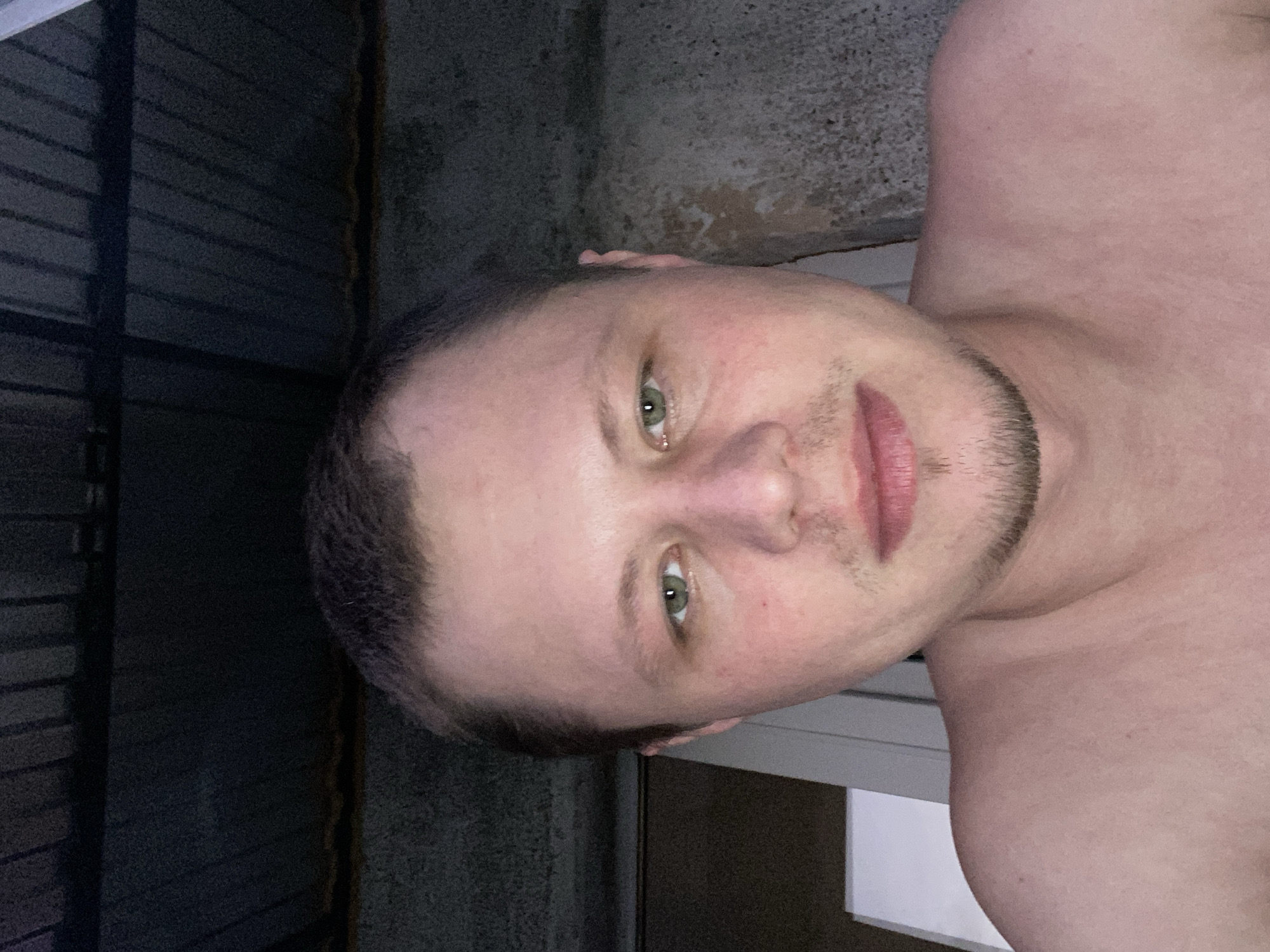 A medium shot shows a Caucasian man in his late 20s or early 30s. He has short brown hair, light green eyes, and a faint goatee and mustache. The man is bare-chested, and his skin appears flushed. He is looking directly at the camera. The background suggests an outdoor setting, possibly a balcony or patio, with a corrugated metal roof overhead and a textured wall to the right. The lighting is dim, indicating it may be evening or night. The image was taken in Beloretsk, Russia.