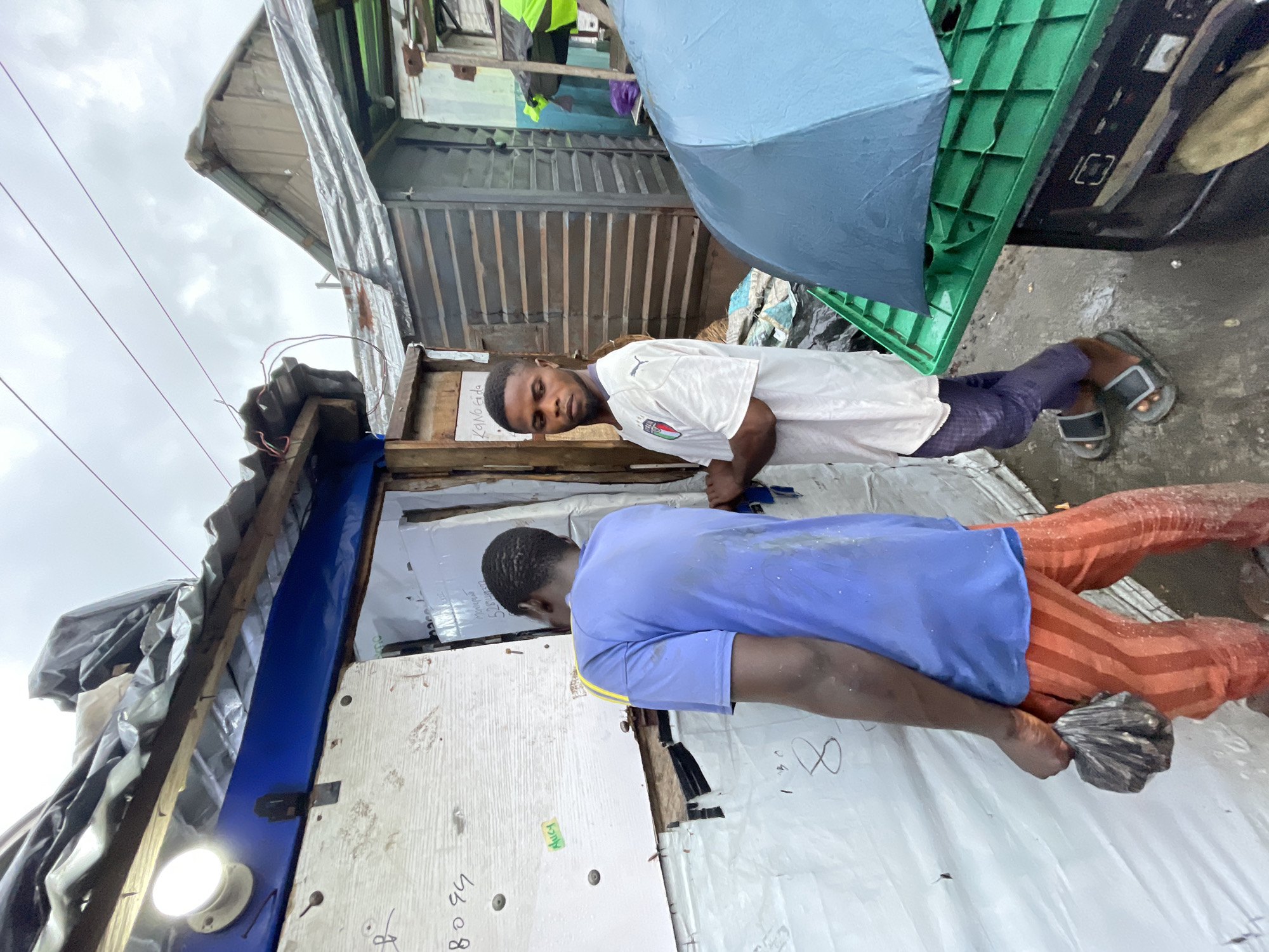 Two young men stand in front of a makeshift structure, likely in a densely populated area of Lagos, Nigeria, on a cloudy day. The structure appears to be constructed from corrugated metal, wood, and plastic sheeting, with a tarp forming the roof. One man, wearing a blue t-shirt and orange striped pants, is facing away from the camera, holding a plastic bag in his right hand. He appears to be wet, as does his clothing. The other man, wearing a white t-shirt with an Italian flag emblem and dark pants, is facing the camera and looking directly at it. He is also standing on wet ground. A large blue umbrella is open and resting on a green plastic crate, partially obscuring a dark object beneath it. Electrical wires are visible overhead. The overall impression is of a basic, possibly temporary living space in an urban environment, with signs of recent rain.