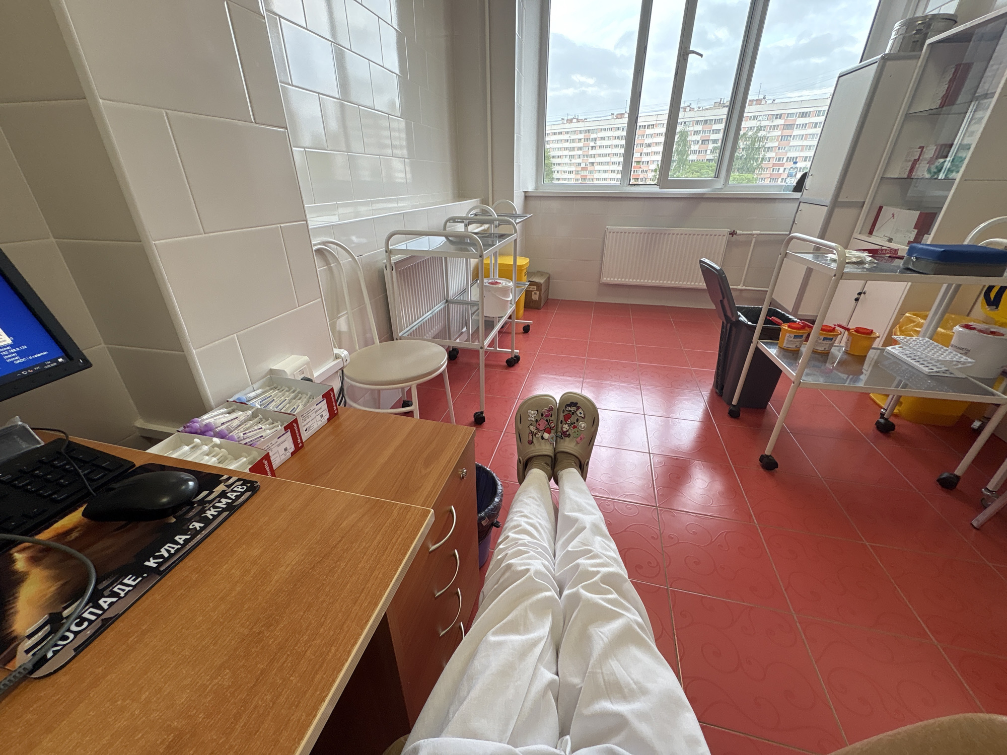 The image shows a person seated at a desk in what appears to be a medical or clinic setting. The person is wearing white pants and Crocs with cartoon embellishments. Their legs are stretched out in front of them, suggesting a relaxed posture.
The environment includes tiled walls, a window with a view of apartment buildings, medical carts and equipment, and a computer workstation. The computer mousepad has Russian text on it.
The setting is likely in Russia, given the text on the mousepad. Based on the architectural style of the apartment buildings visible through the window, the location could be Saint Petersburg, Russia.