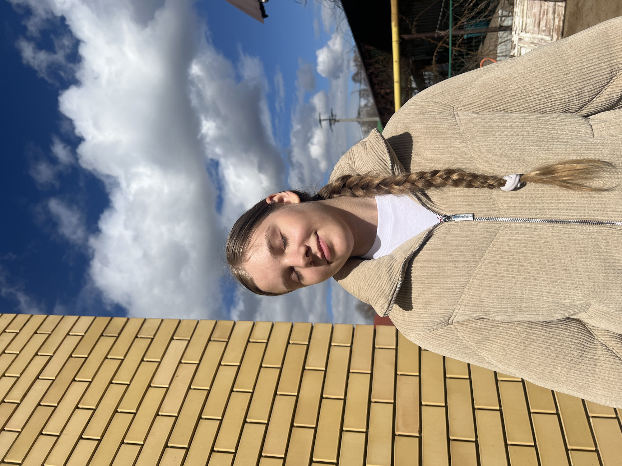 A medium shot captures a young woman, facing forward with her eyes closed in Votkinsk, Russia. She wears a white t-shirt under a zippered, light brown corduroy jacket. Her braided hair, secured with a white tie, falls over her shoulder. The backdrop shows a partly cloudy sky, along with an angled view of a yellow brick wall and, in the distance, trees and a structure with yellow poles.
