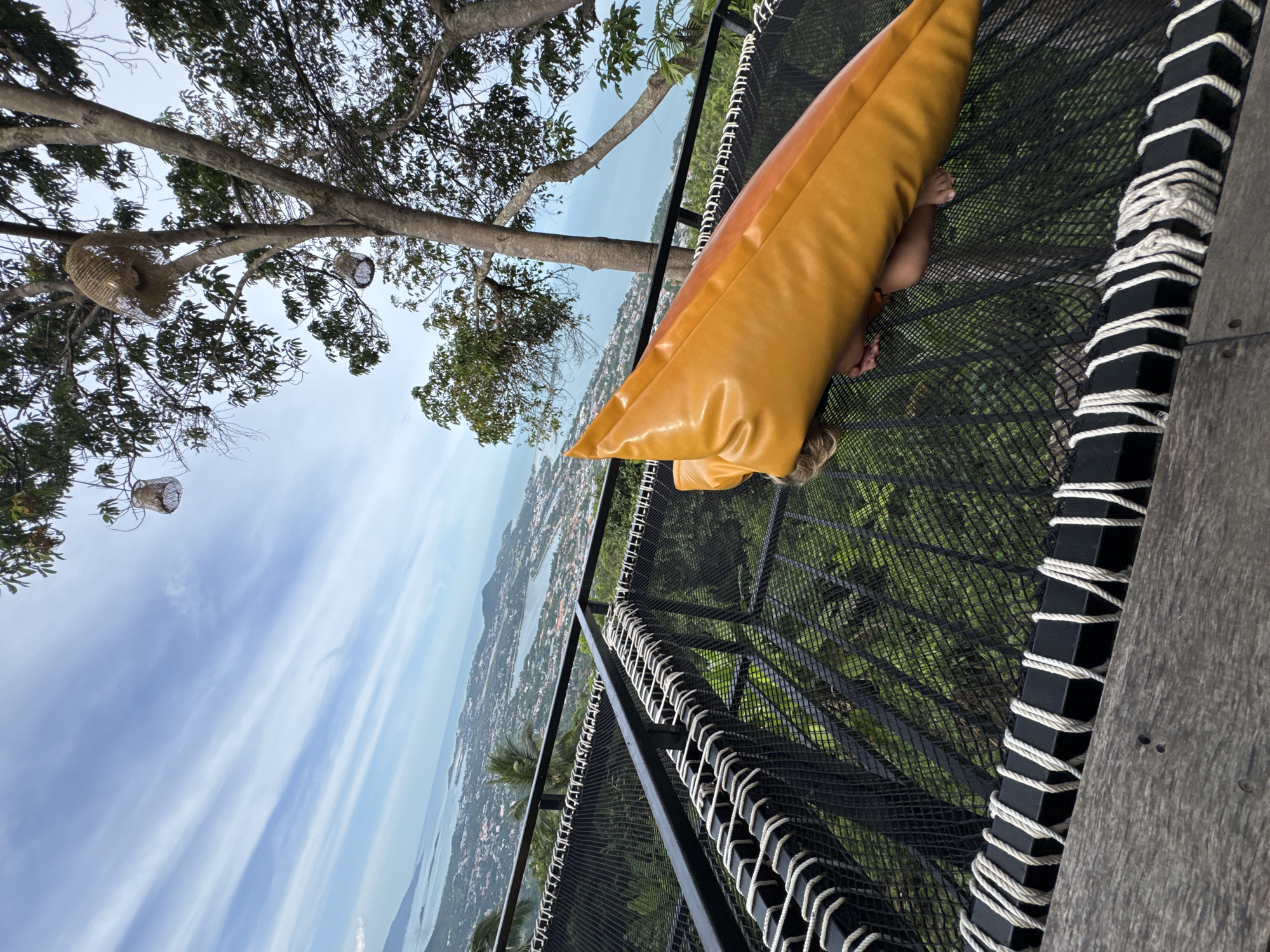 A child is lying on a large, orange beanbag chair on a black net hammock. The hammock is suspended over a green, forested area, and beyond that, a cityscape and ocean are visible under a partly cloudy sky. The hammock is secured to a wooden platform with black metal supports and white rope. There are two woven lamps hanging from a tree branch in the upper portion of the image.