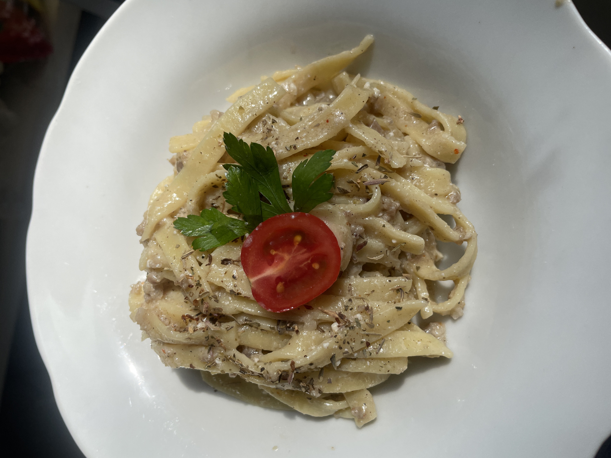 A plate of pasta is presented in a white bowl, garnished with a cherry tomato slice and a sprig of parsley, and sprinkled with dried herbs. The pasta appears to be fettuccine coated in a creamy sauce with visible bits of meat. The lighting suggests an indoor setting, possibly a restaurant or home. The image does not contain any elements that would allow for the identification of a specific location in Başakşehir, Turkey, or any people or actions.