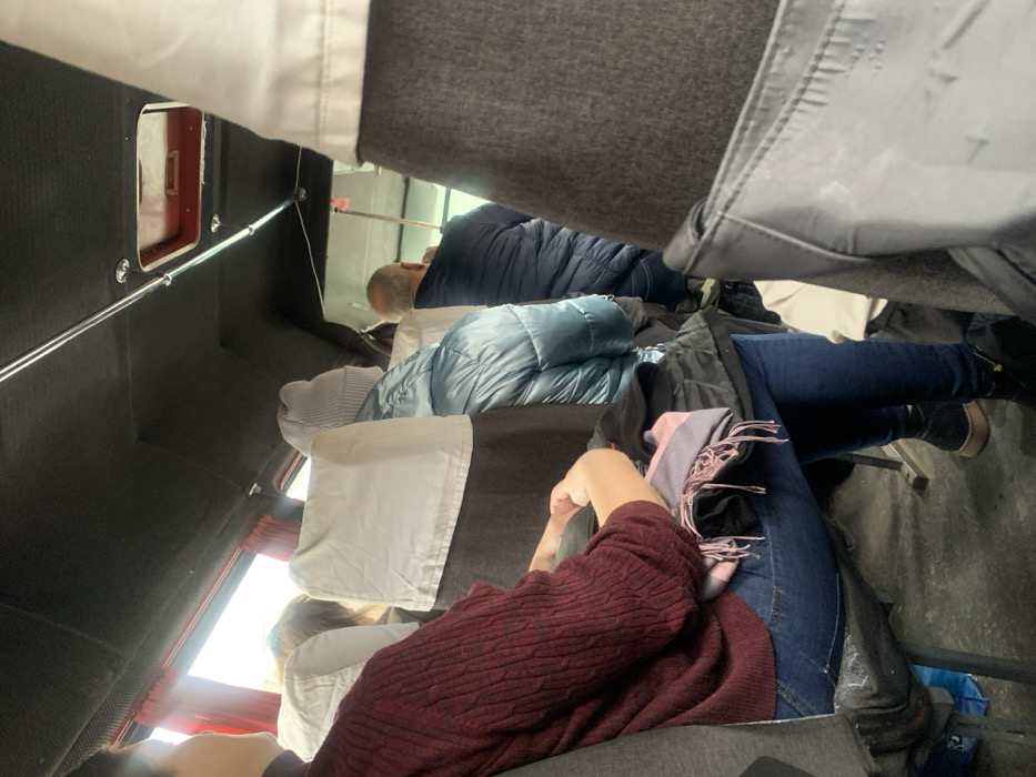 The image displays the interior of a passenger vehicle, likely a bus or large van, located in the city of Kozyn, Ukraine. Multiple individuals are seated, viewed from various perspectives.
In the immediate foreground on the left, a person is seated, wearing a dark red, possibly knitted, long-sleeved top and blue jeans. Their forearms and hands are visible, resting in their lap, where a folded multi-colored scarf with pink, purple, and dark tones, along with other dark fabric items, are placed.
Further into the vehicle, a passenger wearing a light blue puffer jacket and a light grey knitted beanie is seated directly in front of the foreground subject, facing away from the camera. To their right, another individual with short, light-colored hair and wearing a dark blue puffer jacket is also seated with their back to the viewer. Portions of other passengers or seatbacks are visible throughout the interior.
The vehicle's ceiling is dark, featuring exposed metal rails running longitudinally. A rectangular opening with a red frame, consistent with a ventilation or emergency hatch, is visible in the ceiling towards the rear. Windows on the left side are partially visible with reddish-brown curtains or trim, showing bright daylight outside. Seating consists of upholstered chairs, predominantly grey in color, with white fabric covers over the headrests. The floor is dark and indistinct. All visible occupants are in a seated position.