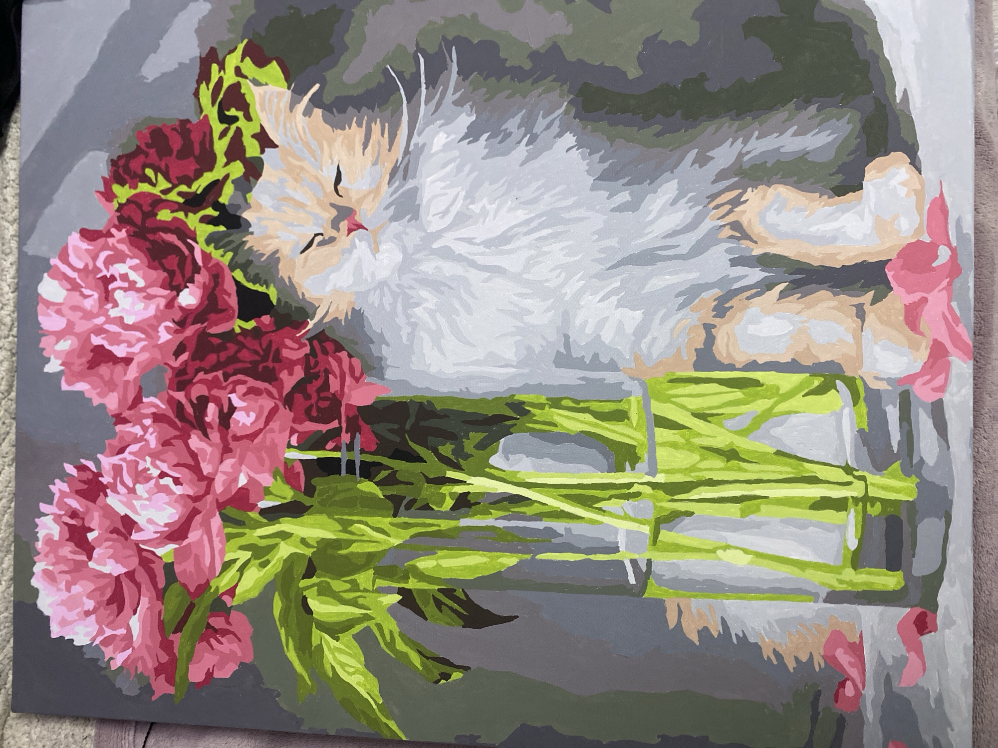 This image is a painting. The painting depicts a fluffy cat sitting next to a vase of pink and red flowers. The cat has its eyes closed and appears to be napping. The flowers are large and vibrant, with green leaves and stems. The vase is made of clear glass and is filled with water. There are also a few fallen flower petals scattered on the surface below the vase and the cat. The background of the painting is a muted gray color, which makes the colors of the flowers and the cat stand out. The painting style is somewhat abstract, with broad brushstrokes and simplified shapes. There are no people or any specific landmarks visible in the painting that would indicate a location in Sochi, Russia.