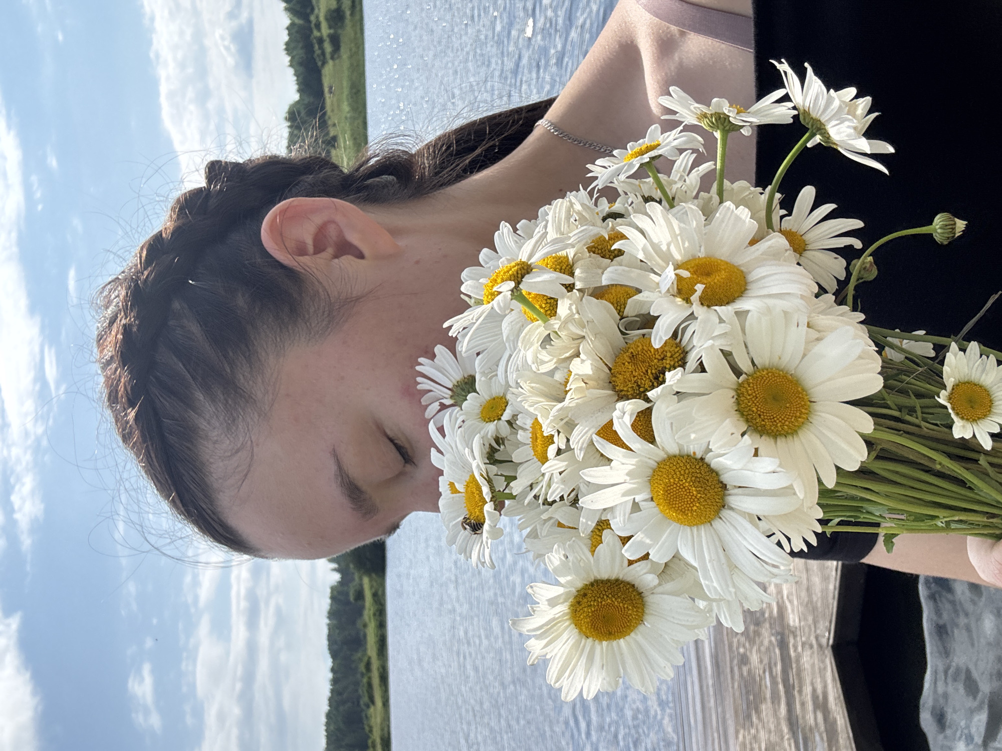 The image shows a close-up of a person's face and a bouquet of daisies, with a body of water and a tree-lined landscape in the background. The person is seen from the side, with their eyes closed and their face tilted downwards towards the flowers. A braid of dark hair is visible, as is a part of their ear and neck. The bouquet is large and full of white daisies with yellow centers. One of the daisies has a bee on it. The person is wearing a black top and a light-colored garment over it. A thin silver chain is visible around their neck.
The scene appears to be outdoors, likely near a body of water, suggested by the reflections and the blue color in the background. The sky is visible with clouds, indicating it is daytime. The overall mood seems peaceful and serene, with the person seemingly enjoying the scent or beauty of the flowers. The location is identified as Pozdyshki, Russia. There is no visible text in the image.
