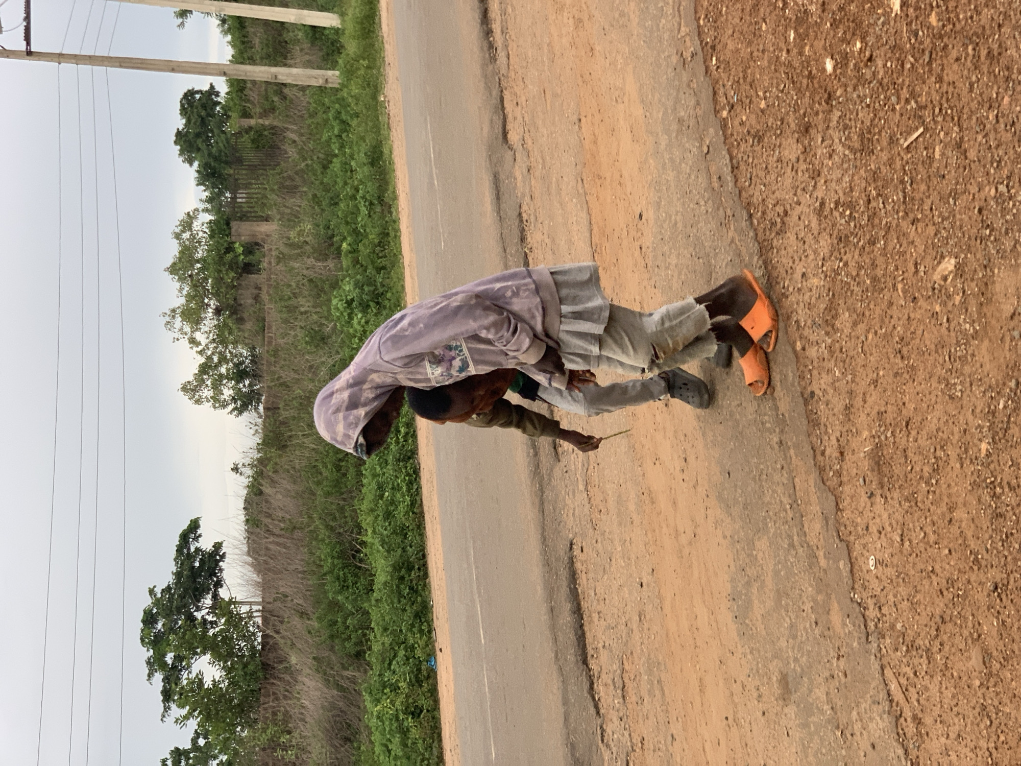 This image captures a roadside scene in Gbeleko, Nigeria. Two individuals, appearing to be children or young adolescents, are positioned on a reddish-brown dirt and gravel shoulder adjacent to a grey asphalt road.
The taller individual, on the right, wears a light purple hooded sweatshirt with a graphic design on the chest, a light grey skirt-like garment over ripped light grey trousers, and orange slide sandals. This person is leaning forward, looking downwards.
The shorter individual, positioned to the left and slightly in front of the taller person, wears a dark-colored top (possibly brown or olive green), light grey trousers, and dark grey Crocs-style shoes. This individual holds a thin, light green stick or plant stem in their right hand, extending it towards the ground, and is also looking downwards. Their face is largely obscured by the taller individual's posture.
The asphalt road, visible in the middle ground, shows signs of wear, cracks, and patches. Beyond the road, on the left, is a bank of green foliage. In the mid-ground, behind the individuals, is an area of dry, light brown grasses and bushes, followed by a row of green trees. Concrete fence posts and what appears to be a metal fence are partially visible within the vegetation in the background. Two concrete utility poles with multiple overhead lines are present on the far right. The sky is overcast with a light grey tone. A small, white circular object lies on the dirt ground in the foreground, and a thin piece of wood debris is also visible.
