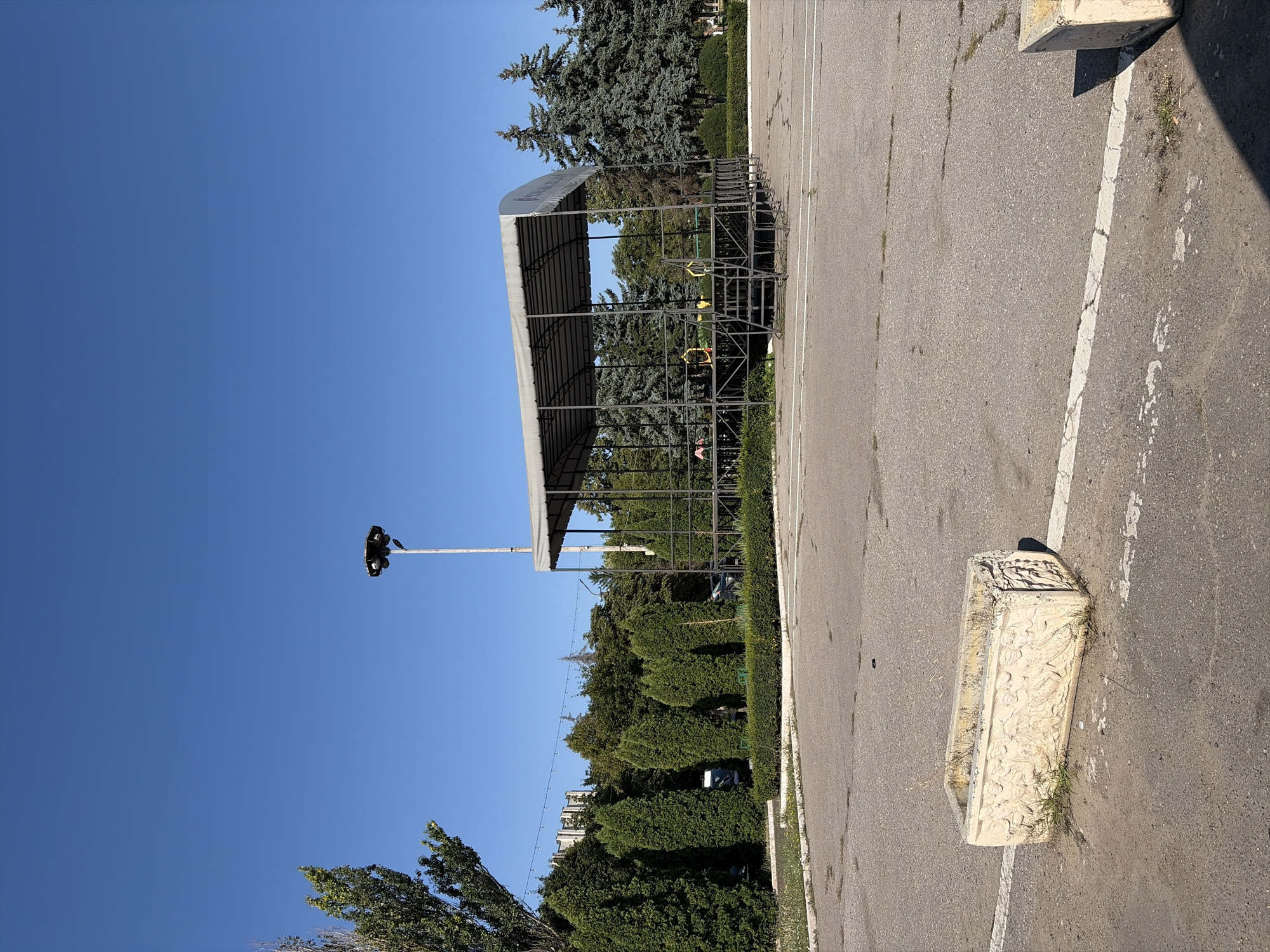 An outdoor asphalt area features a metal stage with a gray canopy. The stage is elevated and has a set of stairs leading up to it. In the foreground, a weathered, rectangular concrete planter with decorative carvings sits on the asphalt. Beyond the stage, a line of tall, conical evergreen trees is visible, followed by a dense backdrop of mixed deciduous and coniferous trees. A tall lamppost with multiple lights stands to the right of the stage, and a clear blue sky fills the upper portion of the image. The scene is devoid of any people or ongoing activity, suggesting a quiet, possibly preparatory, setting. The overall environment appears to be a park or public square area within the city of Criuleni, Moldova.