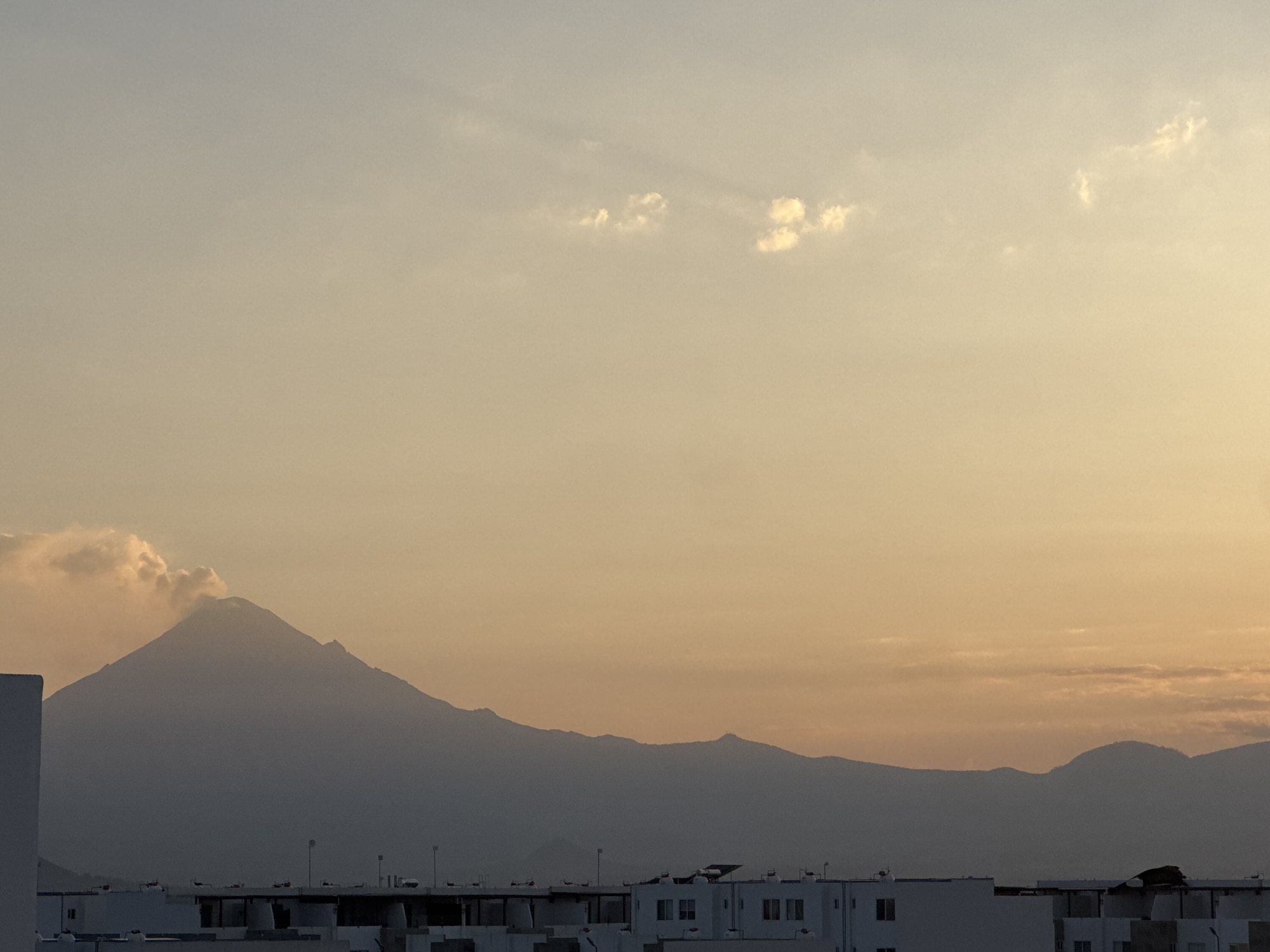 This outdoor image presents a breathtaking landscape featuring a prominent mountain range silhouetted against a vast, gradient sky. The time of day appears to be either sunrise or sunset, indicated by the soft, diffused light, with the sky transitioning from warm, golden-orange tones near the horizon to cooler, lighter blue-grey shades higher up. Scattered, wispy clouds are present, with some catching the light.
The central subject is a majestic mountain range stretching across the middle ground. The most striking feature is the tall, conical peak on the left, which strongly resembles a volcano, with a visible plume of smoke or cloud gently rising from its summit. The mountains are rendered in deep grey and blue-grey silhouettes due to the backlighting and atmospheric haze, revealing their varied contours.
In the foreground, the rooftops of numerous buildings are visible, suggesting an urban or suburban setting. These structures are predominantly light-colored, likely white or off-white, with flat roofs. Details such as windows, structural elements, and what appear to be small installations or water tanks are discernible on the rooftops. The architectural style hints at residential or commercial buildings.
The scene conveys a sense of tranquility and natural grandeur, with the imposing volcano dominating the horizon above the human habitations. The primary activity is the scenic display of the natural environment, possibly highlighting minor volcanic activity. Given the context of San Francisco Ocotlán, Mexico, the volcano is likely Popocatépetl. There is no visible text in the image.