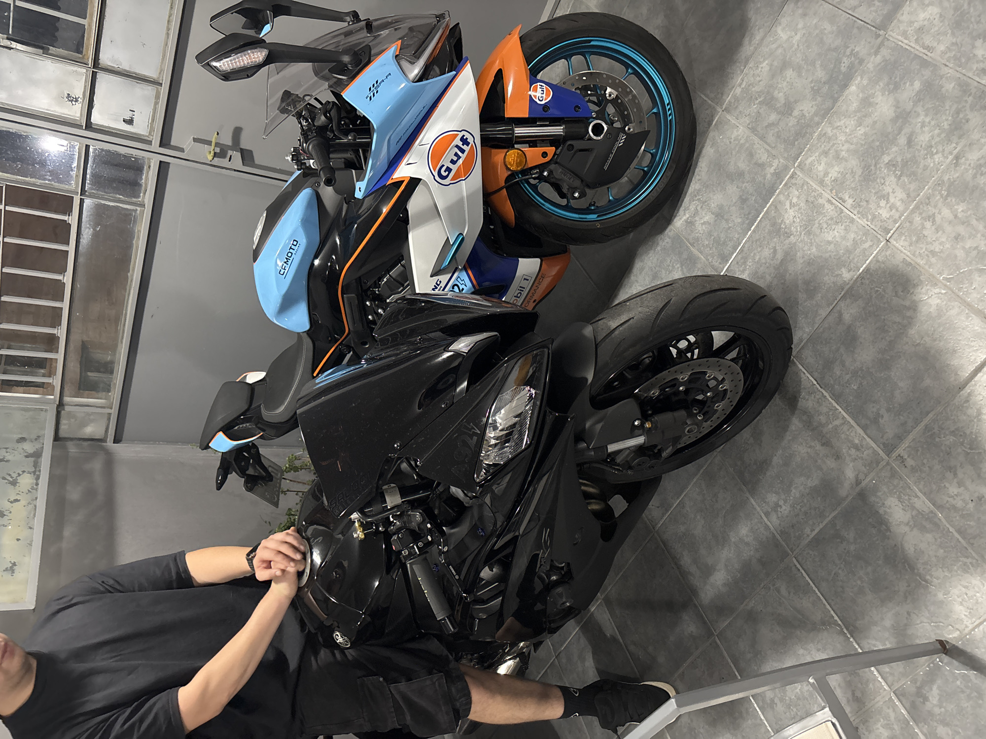 The image shows two motorcycles parked indoors. The one in the foreground is a black motorcycle with its front wheel facing the viewer. A person wearing a black t-shirt, black shorts, and black shoes is standing partially in front of the black motorcycle, with their hands resting on the fuel tank. Behind the black motorcycle is another motorcycle with a distinctive Gulf Oil livery (orange, white, and blue). The environment appears to be a garage or storage space with tiled floors and windows in the background. Lettering such as "CFMOTO", "NG25", "Mobil 1", and "Gulf" can be seen on the motorcycles. There is no information in the image to verify that the location is in the city of Cordoba, Argentina.