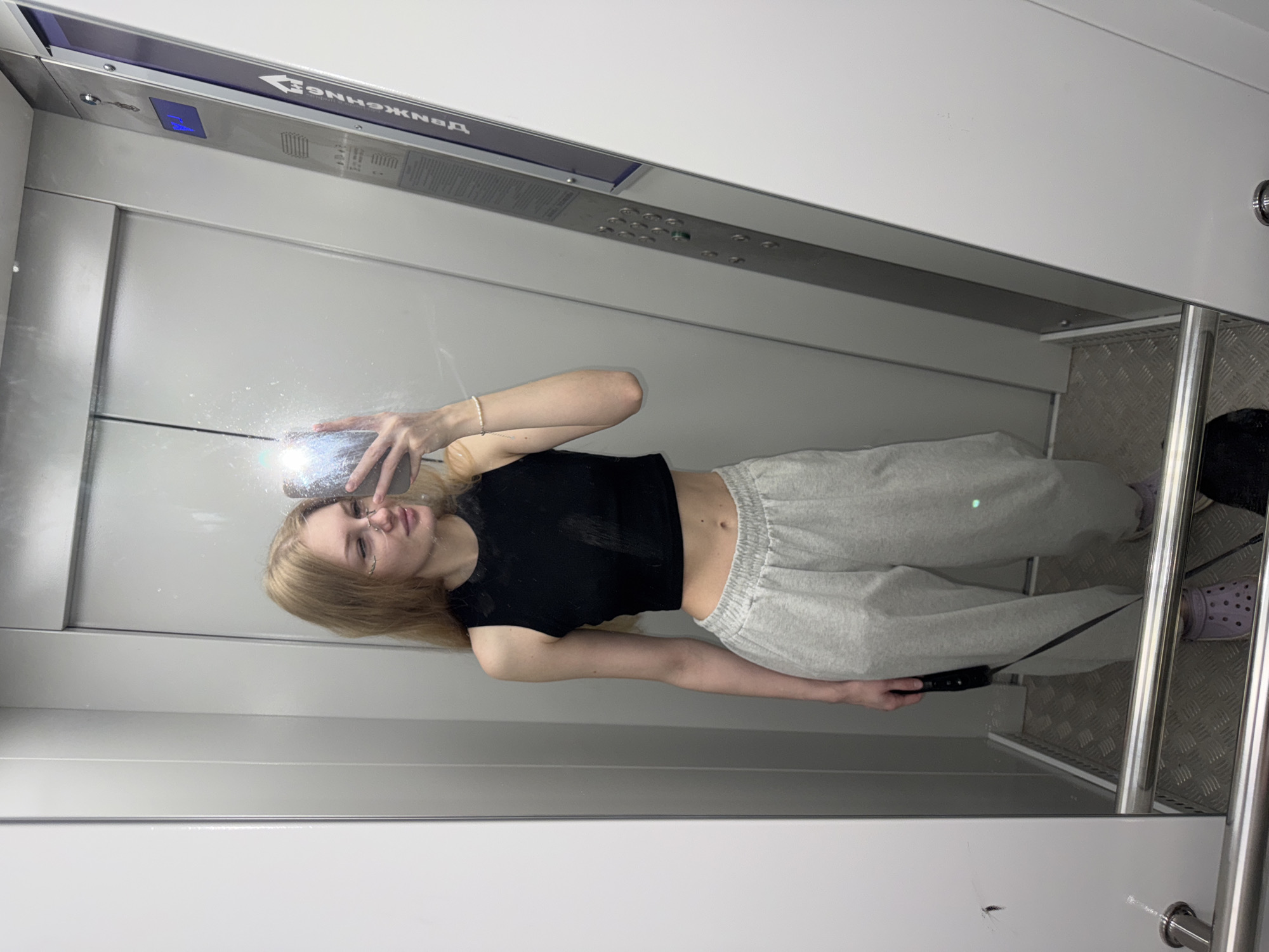 A young woman with blonde hair and glasses is standing in an elevator, taking a mirror selfie. She is wearing a black cropped tank top and light gray sweatpants. The elevator has gray walls and a metal floor. A metal railing runs across the bottom of the frame. On the right side of the elevator is a control panel with buttons and a digital display. The elevator appears to be in a residential building in the city of Perm, Russia, as indicated by the Cyrillic text on the control panel, which reads "ДВИЖЕНИЕ" (movement).