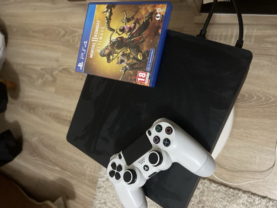 An indoor scene depicts a black PlayStation 4 (PS4) console positioned on a partially visible white base. A white DualShock 4 controller rests on the left surface of the console, with a white cable extending from its front, trailing onto a light-colored shaggy rug visible on the bottom left. To the right of the console, a blue PlayStation 4 game case for "Mortal Kombat 11 Ultimate" is displayed, featuring an "18" age rating logo. Two black cables are connected to the rear-right of the console, extending out of frame towards the bottom right. The floor consists of light-toned wooden laminate. The background shows blurry indications of furniture. No individuals are present, and no actions or interactions are occurring. The specific location within Sedlartsi, Bulgaria, is not discernible from the visual evidence provided.