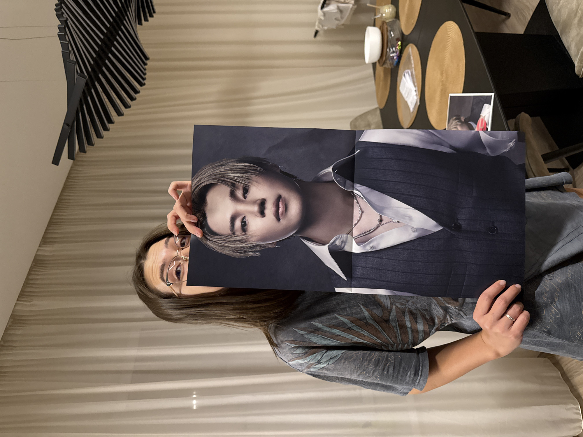 A woman with brown hair and glasses holds up a large portrait of a young Asian man in front of her face, partially obscuring her features. The man in the portrait has light brown hair and wears a white collared shirt with a dark vest. The background of the portrait is a dark gray gradient. The woman is wearing a gray shirt with a pattern. The setting appears to be indoors, possibly in a dining area. A black table with place settings and decorative items is visible to the right, along with a chair. A modern, geometric black art piece hangs on the wall to the left. The exact location in Krasnoyarsk, Russia, is not discernible from the image.
