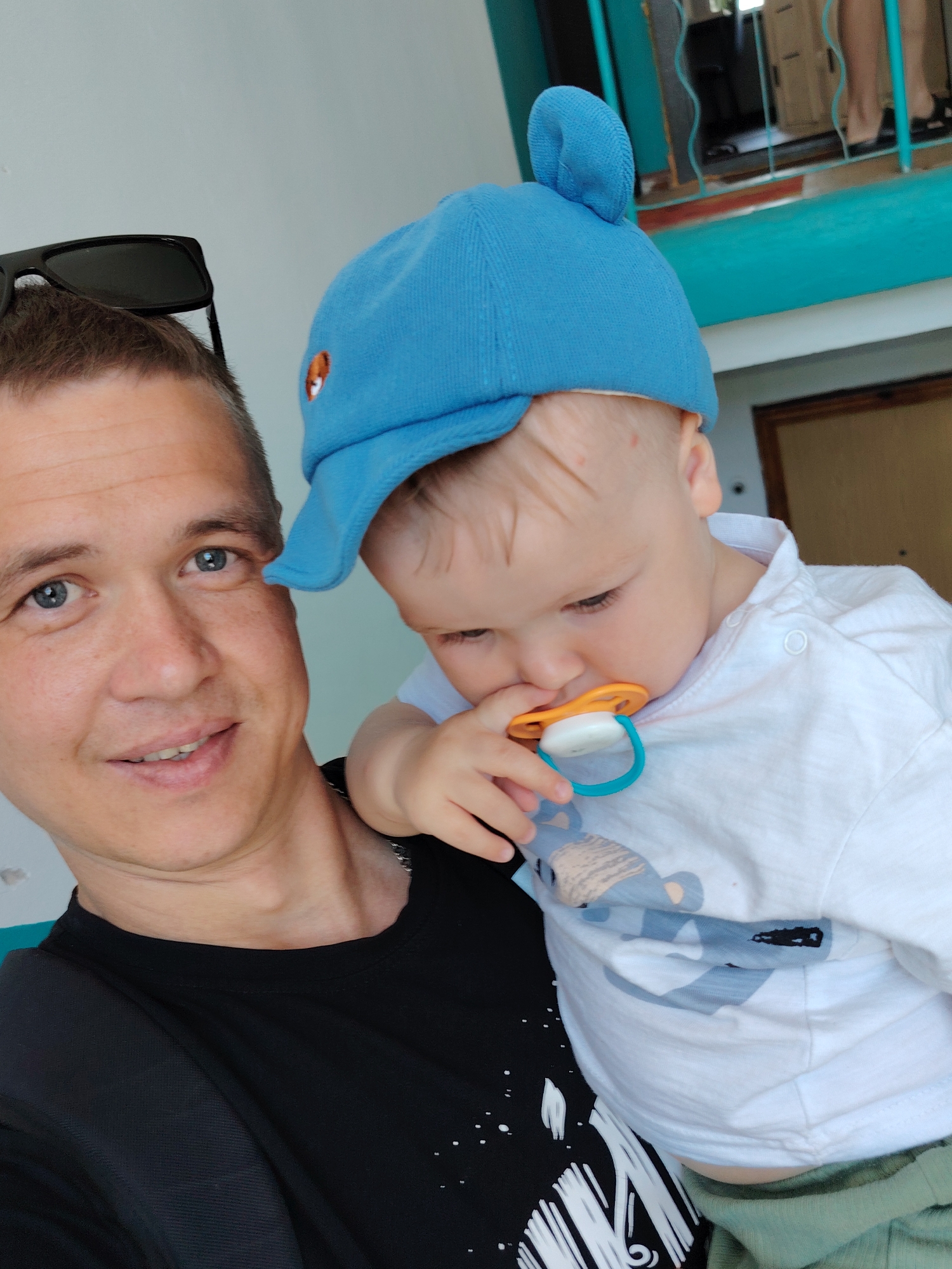 A man with short, graying hair and blue eyes is holding a baby in his arms. The man is wearing a black t-shirt and has sunglasses perched on his head. The baby is wearing a blue cap and a white t-shirt with a light blue bear graphic. The baby has a pacifier in its mouth. The background includes a turquoise painted structure with window frames. The scene appears to be in Chaikovsky, Russia.