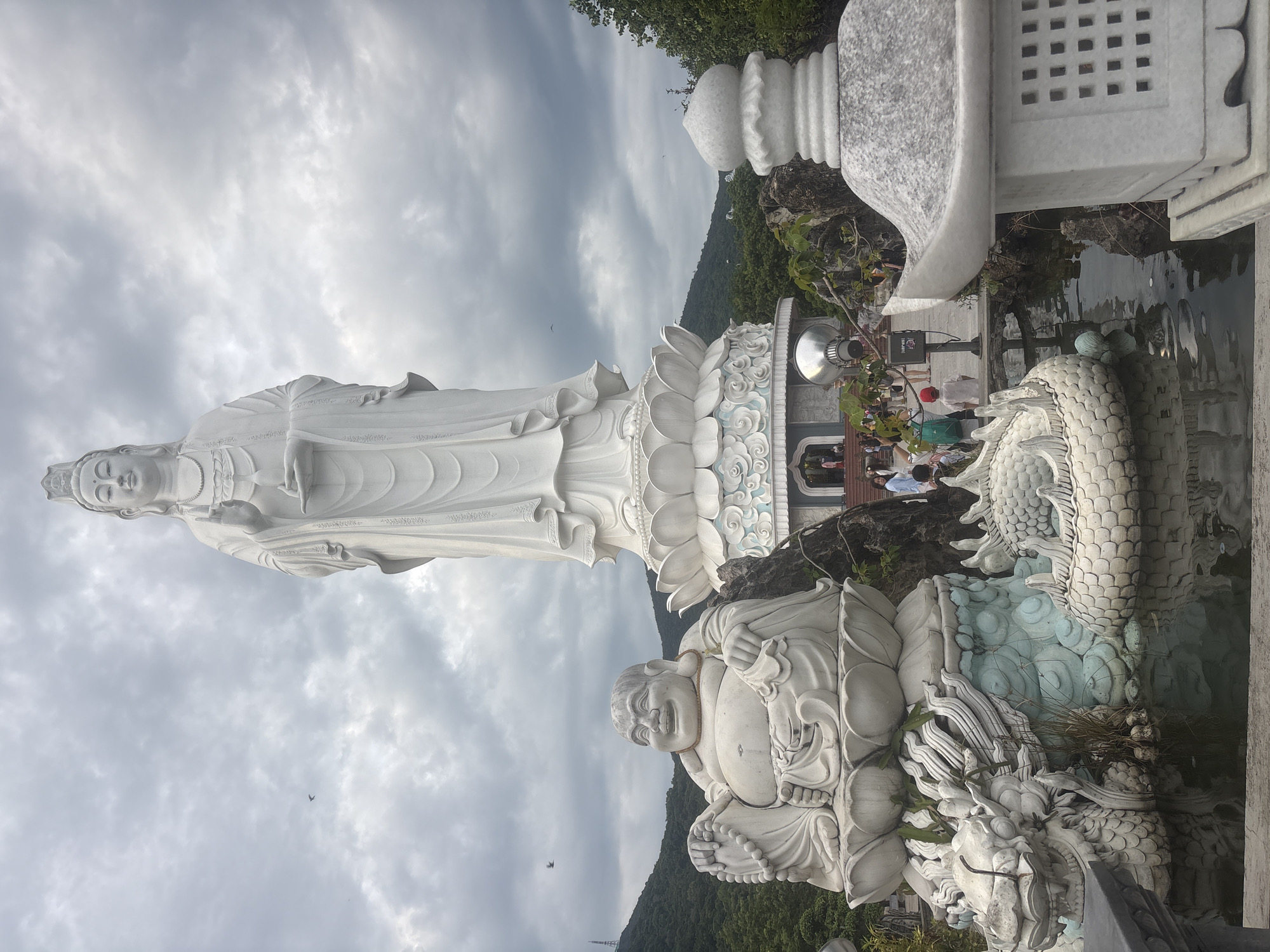 The image shows a large white statue of a goddess, likely Guan Yin, with a serene expression and adorned in traditional robes. She is standing on a lotus flower pedestal, which is part of a larger monument. In the foreground, to the left of the Guan Yin statue, is a smaller statue of a laughing Buddha, also made of white stone. This Buddha statue is seated and has a cheerful demeanor, with prayer beads draped around its neck.
Both statues are positioned near a body of water, possibly a pond or a reflection pool, where the dragon sculptures are visible. The dragon sculptures are white and intricately detailed, coiled around the base of the Guan Yin statue and appearing to emerge from the water. In the middle ground, there are several people, dressed in casual attire, walking on a pathway. Some are looking at the statues, while others are engaged in conversation.
The background features lush green hills and a cloudy sky, suggesting an outdoor setting. To the right of the Guan Yin statue and in the foreground, a white stone lantern is visible, characteristic of East Asian gardens. This scene is located at the Linh Ung Pagoda on Son Tra Peninsula in Da Nang, Vietnam. The pagoda is a significant Buddhist site known for its large statues and scenic views.