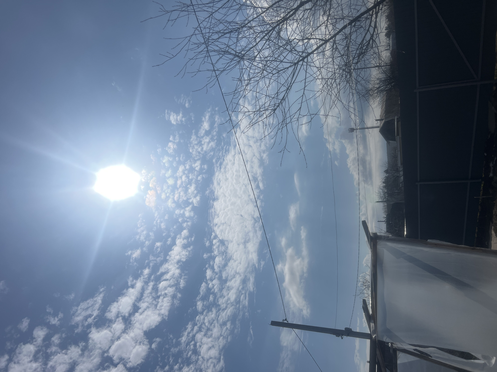 This image, taken in Chaikovsky, Russia, captures a bright outdoor scene dominated by a clear blue sky with scattered white cumulus and cirrocumulus clouds. The sun is a powerful, intensely bright light source positioned in the upper left, casting significant lens flare.
Bare tree branches, indicative of late autumn, winter, or early spring, are silhouetted against the sky in the upper right corner and along the distant horizon. Several dark utility wires crisscross the frame, with one prominent wire stretching diagonally from the upper left to the lower right. A dark, wooden utility pole stands in the lower center-right, supporting these wires.
In the lower right portion of the image, there is a structure, likely a greenhouse or a simple shelter, constructed with wooden supports and covered in translucent white plastic sheeting. Adjacent to it, on the far right, a dark, possibly metal-framed structure or covered area is partially visible. The background reveals silhouettes of distant buildings or structures along a hazy horizon.
The scene depicts a static moment with no explicit activity taking place. The time of day appears to be mid-day due to the high position and intensity of the sun. There is no visible text in the image.