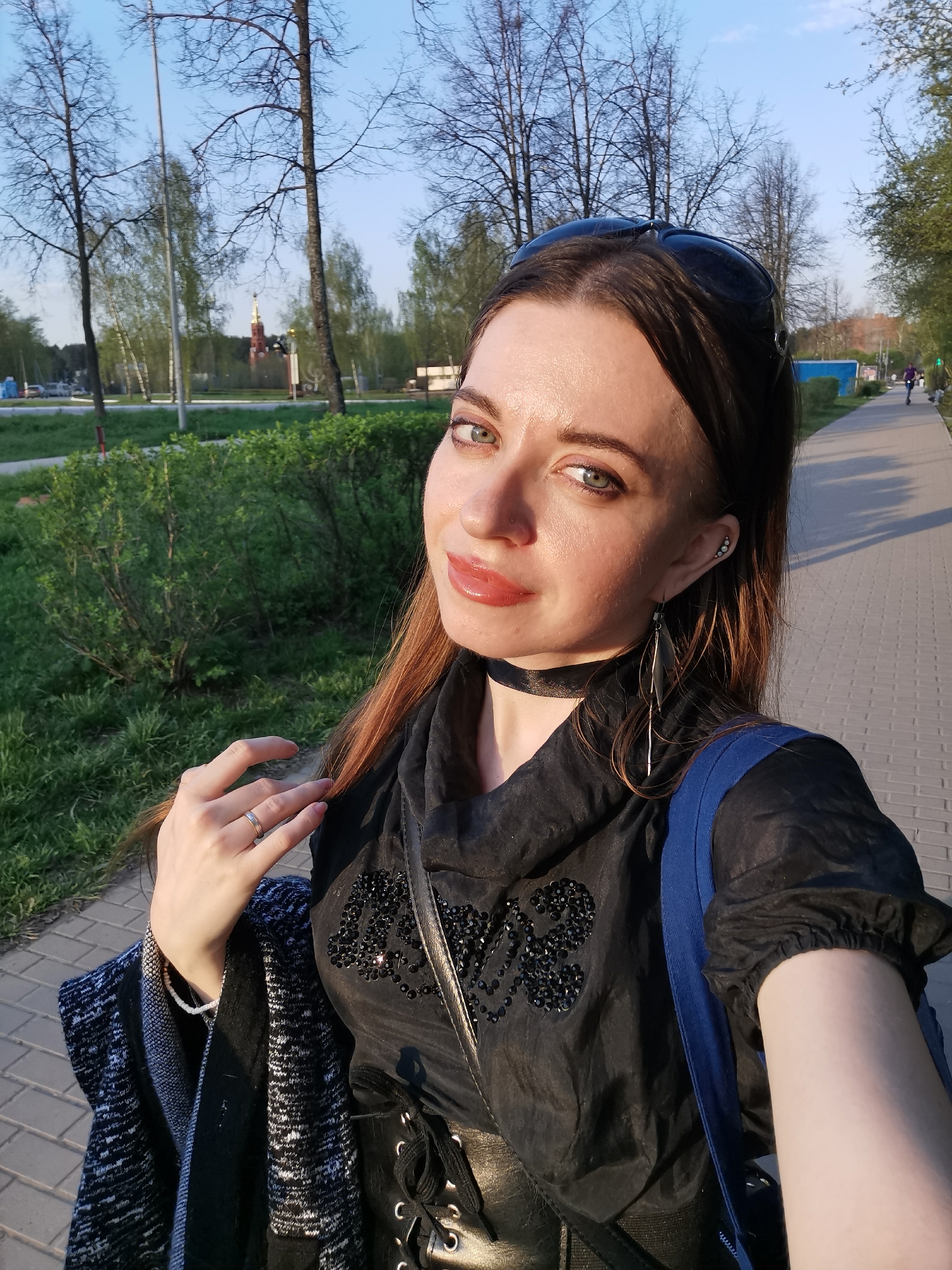 A woman is taking a selfie outdoors on a paved walkway. She has medium-length brown hair, green eyes, and fair skin. She is wearing a black choker, long earrings, a black top with embellishments, and a possible corset-like belt. Sunglasses are perched on her head. She has a blue backpack slung over her shoulder and is holding a patterned shawl or jacket in one hand.
The background shows a park-like setting with green bushes, trees, and a paved pathway. In the distance, there is a building with a steeple. The location appears to be in the city of Unknown, Unknown. Another person is walking along the path in the distance. The lighting suggests it is late afternoon or early evening.