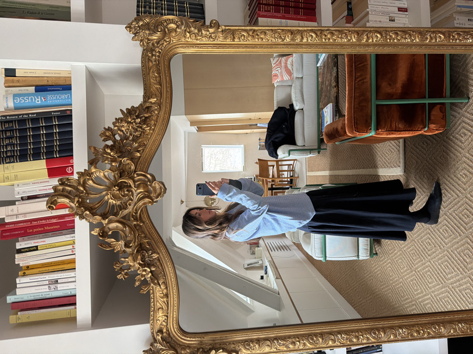 The image displays a young adult female taking a mirror selfie within an apartment in Paris, France. The subject, with shoulder-length brown hair, is seen from the chest down, wearing a light blue and white striped long-sleeved shirt, a long black skirt, black tights, and black flat shoes. She holds a dark-colored smartphone with both hands, angled to capture her reflection in a large, ornate gold-framed mirror that dominates the foreground.
The apartment's interior, visible in the mirror's reflection, includes a living area and a dining space. The living area features a white sofa and a burnt orange velvet armchair, both distinguished by green metal frames, positioned on a light brown geometric-patterned rug. A dark jacket is draped over the white sofa, and a small coffee table with books rests in front of it. In the background, a dining table with several wooden chairs is discernible. To the left, a sloped ceiling with two rectangular windows and a long, white built-in cabinet indicate an attic or mansard-style dwelling.
Above the mirror, white shelves are densely packed with books, many with French titles such as "Larousse," "Russe," and "Linguistique." Additional bookshelves with red and black bound volumes are partially visible to the right of the mirror's frame.