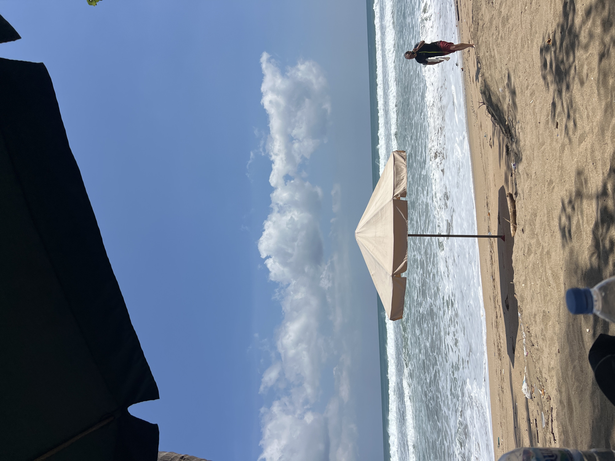 A bright, sunny day at Kuta Beach in Indonesia, with a person walking on the sand near the ocean. The beach is sandy and there is a large umbrella providing shade. The ocean has moderate waves and the sky is clear blue with some clouds. The person is wearing a dark shirt and red shorts and is carrying a surfboard. There are some plastic bottles and debris on the sand.