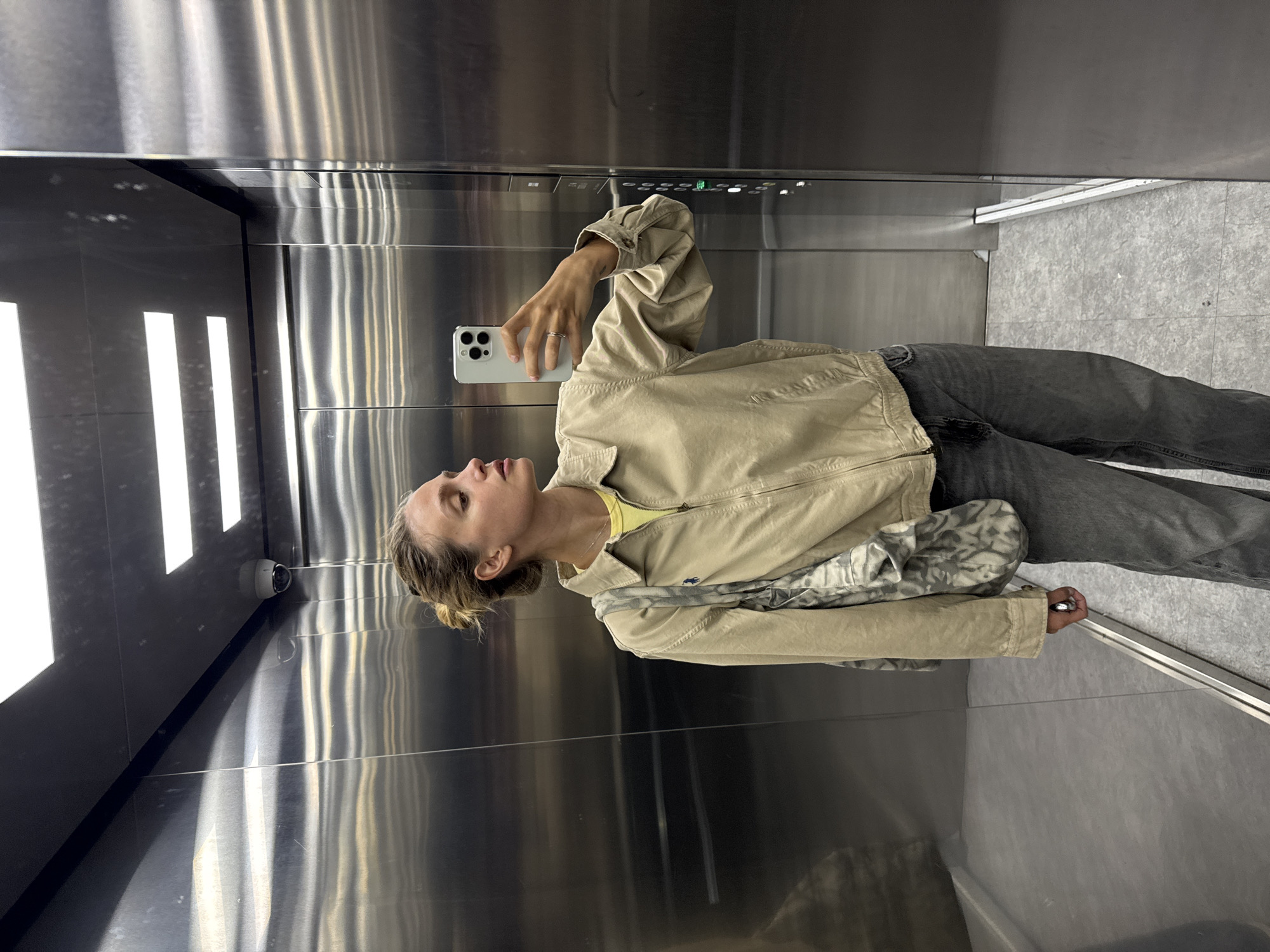A woman is taking a selfie in an elevator. The elevator is made of stainless steel and has recessed lighting on the left side. There is a security camera mounted on the wall. The woman is wearing a tan jacket, a yellow shirt, and gray jeans. She is holding a white smartphone in her right hand. The floor of the elevator is gray tile.