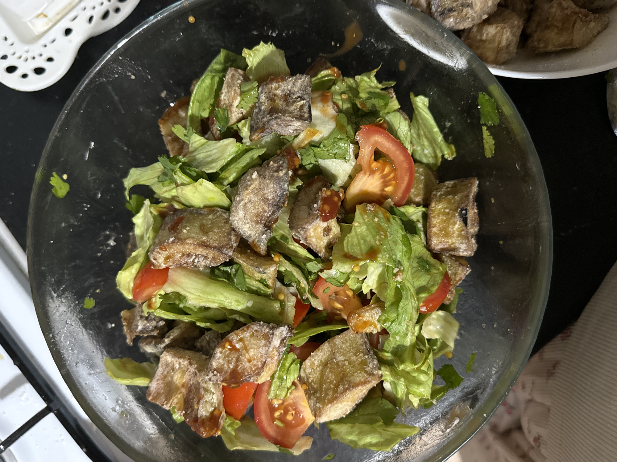 The image shows a bowl of salad. The salad contains lettuce, sliced tomatoes, and fried tofu cubes. The bowl is glass and sits on a surface near a stove. Another plate with similar food items is also visible. The location in Chaikovsky, Russia is not determinable from the image.