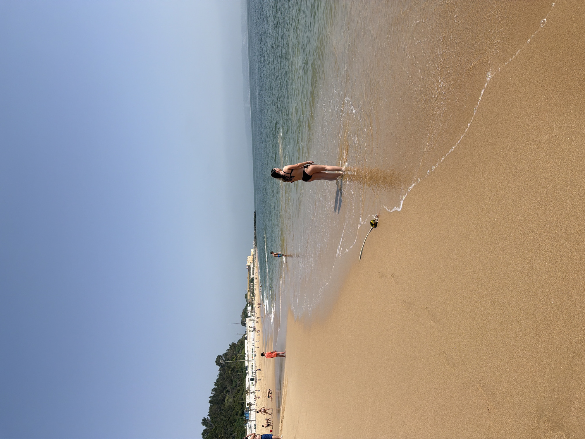 A horizontal wide shot captures a beach scene in Oeiras, Portugal, on a bright, sunny day. The foreground features a large expanse of tan sand, receding towards the middle ground where the sea meets the shore. A woman stands waist-deep in the water, facing away from the camera and towards the ocean. She is wearing a black bikini. Further down the beach, other people can be seen walking and standing, their figures becoming smaller with distance. In the background, a line of buildings and trees is visible along the coastline. The sky above is clear and blue. The sea is calm, and a light wave is breaking near the woman's feet. The entire scene is bathed in sunlight, creating a warm and inviting atmosphere.