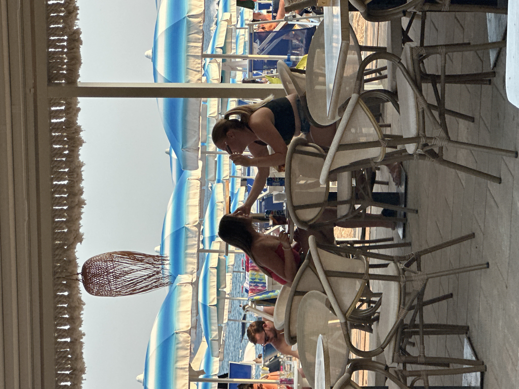 The image depicts a beachside cafe with patrons seated at tables. Two women are the primary subjects in the foreground, positioned at a table. The woman on the left, wearing a red top, is looking towards the woman on the right. The woman on the right, wearing a black top, is applying makeup to her face. Several other tables and chairs are visible in the cafe setting. Behind the cafe, a beach with numerous blue and white striped umbrellas and people relaxing can be seen. The sea is visible in the background, with a clear sky above. The overall scene suggests a casual, outdoor dining experience at a coastal location. The location is identified as Boreti, Montenegro.