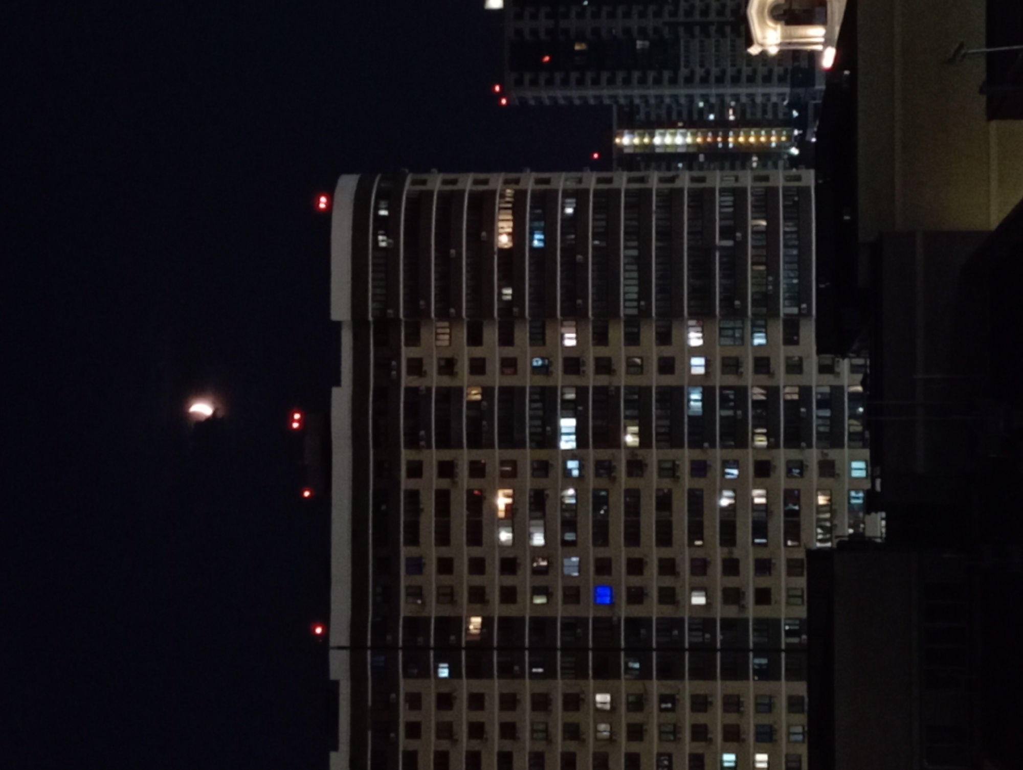 The image depicts a night scene in the city of Krasnodar, Russia, featuring a large apartment building and other structures against a dark sky with a crescent moon. The apartment building, with its illuminated windows, dominates the center of the frame. The windows are lit in various shades of yellow and white, suggesting occupied apartments. A single window on the building's facade glows with a distinct blue light. Red lights are visible on the top of the apartment building and other taller structures in the background. These red lights are likely navigational warning lights for aircraft. The overall impression is that of a densely populated urban environment at night.