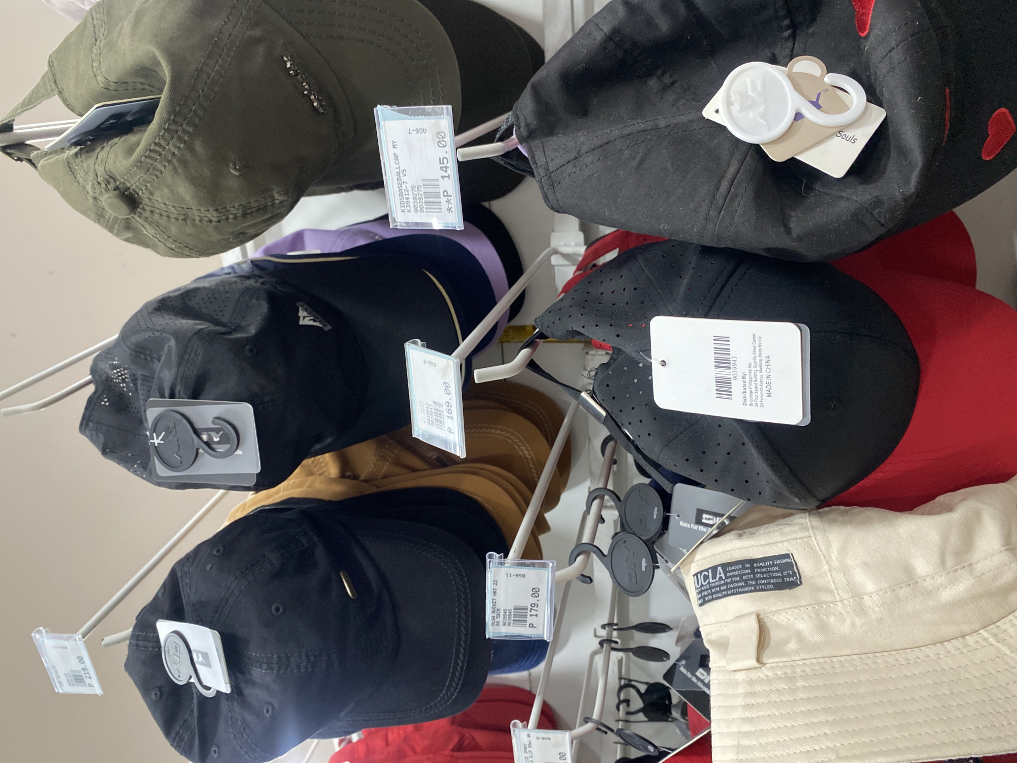 The image captures an overhead view of various hats displayed for sale on white, metal retail hooks, likely in a store in Davao City, Philippines. The display features a range of headwear, predominantly baseball caps and bucket hats, in different colors and styles.
In the upper left, an olive green baseball cap is visible, partially obscuring a price tag. Below it, several baseball caps hang in a row: one black with what appears to be a purple or lavender underside, another black cap with a prominent logo, and a tan/brown cap. On the right side, there's a black baseball cap with red embroidered accents. Below it, a black perforated sports-style cap is displayed next to a red cap. At the bottom right, a beige or cream-colored bucket hat with visible stitching is positioned. Some caps feature small, dark anti-theft tags.
Visible price tags and product information include:
* A tag showing "**P 145.00**" for a "KIDSBASEBALLCAP MY K39412-7 VS".
* Another tag indicates "**P 169.00**".
* A tag displays "**P 179.00**" for a "MESH BUCKET HAT 22".
* A tag showing "**P 219.00**".
* Manufacturer details on a tag read: "Distributed By: Bricolage Philippines Inc. 3A Floor Xeland Building, Guerilla Street Corner Gil Fernando Avenue, Marikina, Metro Manila MADE IN CHINA."
* A white tag on a black cap shows "Souls" and a stylized logo.
* The beige bucket hat has a black fabric label stitched onto it with the text: "UCLA LEADER IN QUALITY FASHION IMPRESSION. FUNCTION. ADVANCE FASHION FOR YOU. BEST SELECTION. IT'S ABOUT OUR PASSION. THE CONFIDENCE THAT BRINGS QUALITY OUTSTANDING STYLE."
* Another partially visible tag reads "Hats for the".
The overall scene suggests a typical retail environment showcasing accessories. No people are visible.