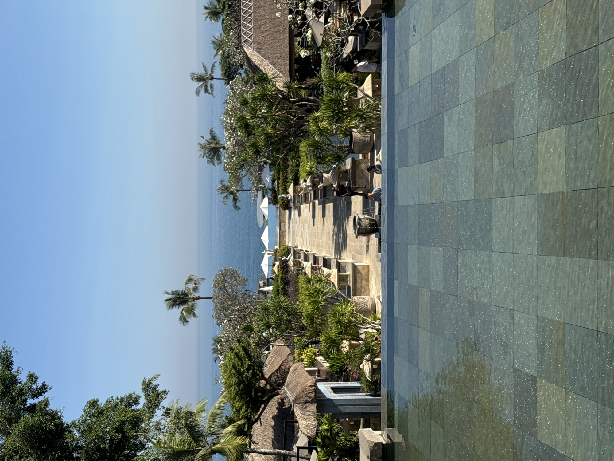 The image depicts a seaside resort in Kuta Selatan, Indonesia, captured from an elevated perspective, likely from a balcony or rooftop area. The scene blends natural and man-made elements.
In the foreground, there's a stone tiled surface that seems to be wet, possibly from a recent cleaning or rain. This area transitions into a walkway lined with decorative stone planters, tropical trees, and shrubs. The walkway leads towards the ocean, where white umbrellas are visible on the beach.
The ocean appears calm and merges with a clear, blue sky. Several palm trees are dotted along the shoreline and throughout the resort, adding to the tropical ambience.
Thatch-roofed structures are also visible, possibly part of the resort's buildings. Several people are visible along the walkway.