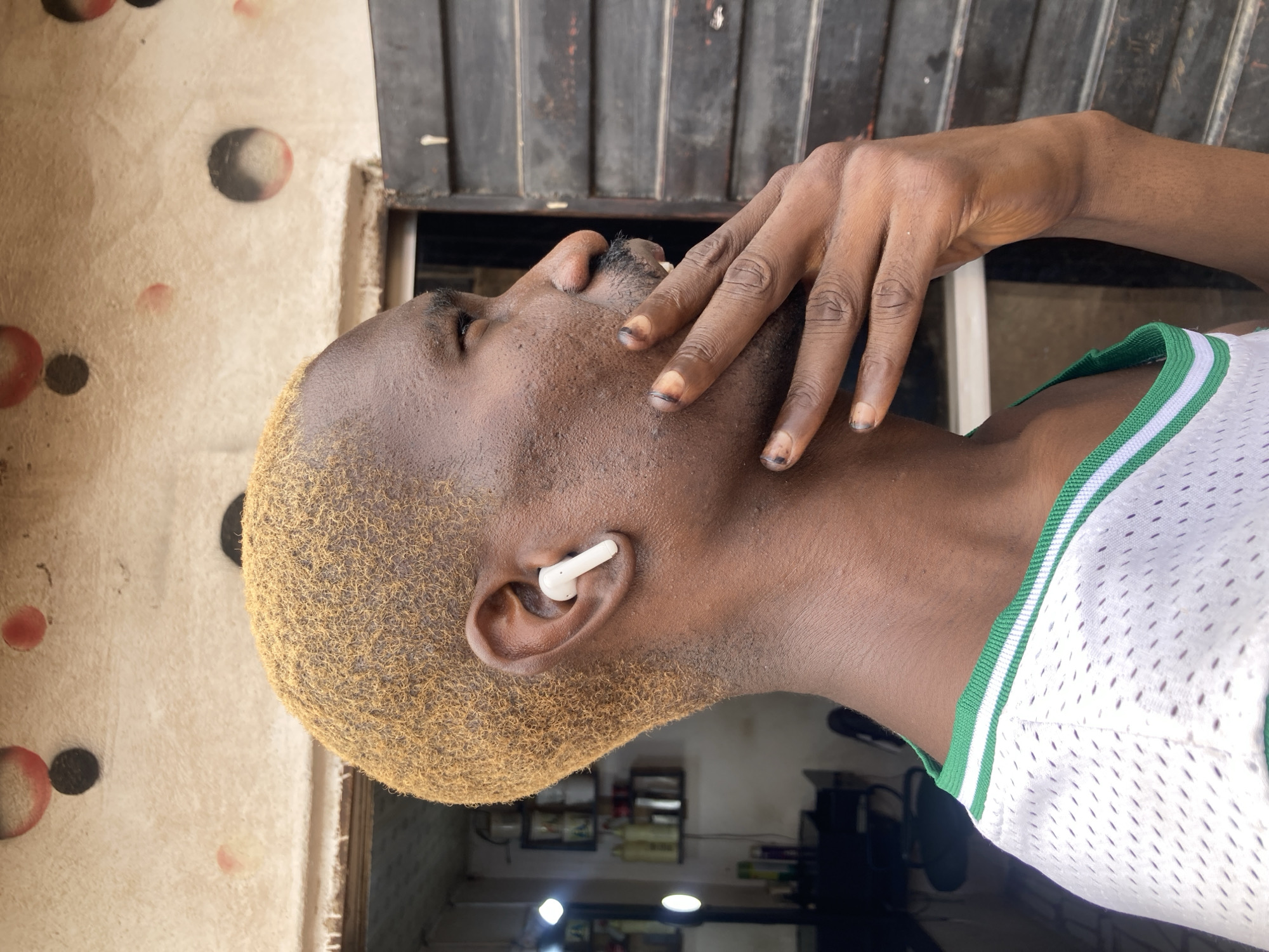 A medium close-up shot from the side shows a Black man in Gwong, Nigeria, with short, bleached blonde hair. He is wearing a white jersey with green trim. He has a white wireless earbud in his right ear and is touching his chin with his left hand. His fingernails appear to have dark polish or dirt on them. In the background, a building with a textured, light-colored wall decorated with red and black circles is visible. To the right of the man, a dark, slatted window or door is present. To the left, a blurry interior with shelves of bottles and a lit mirror suggests a salon or shop.