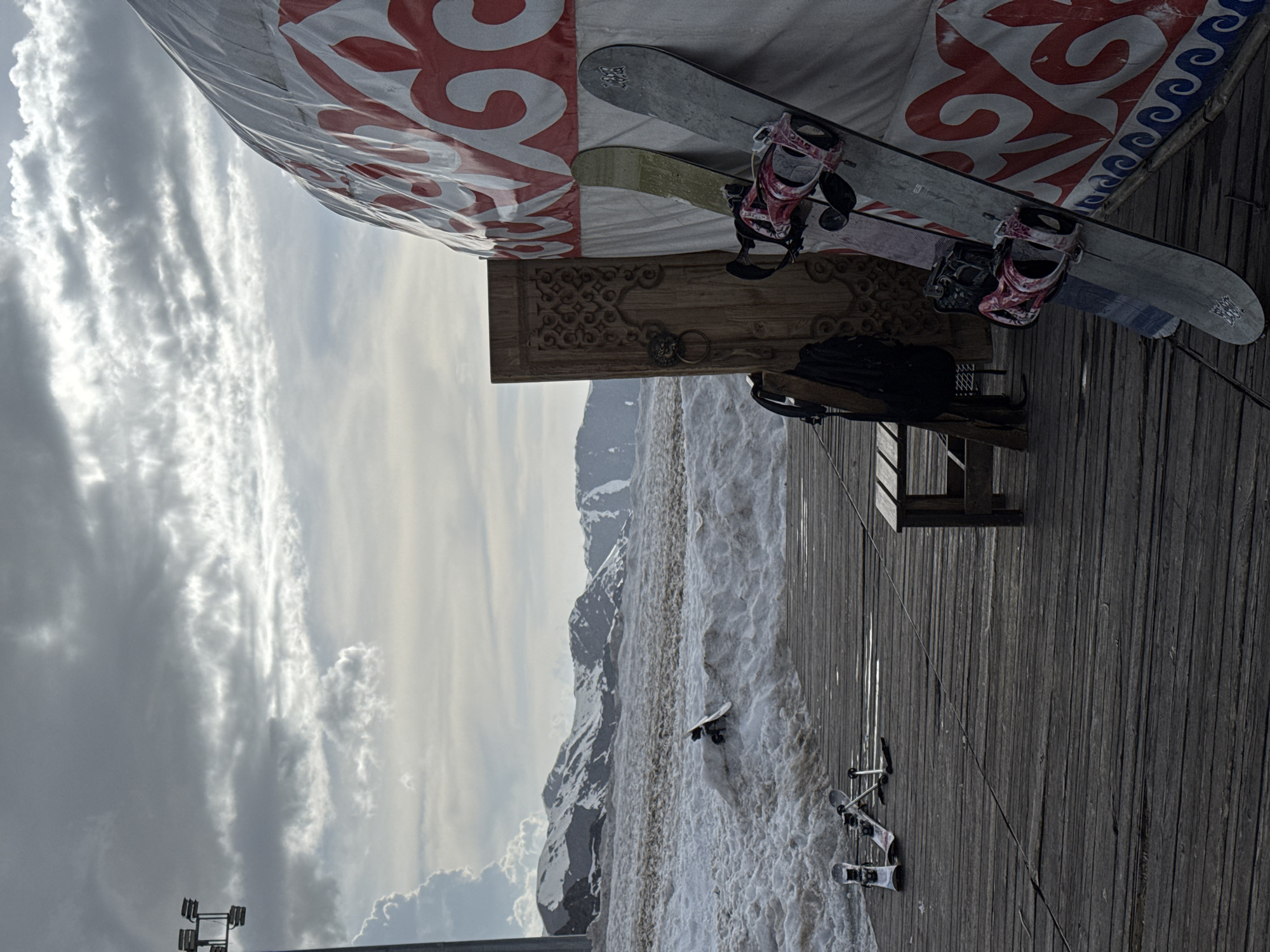 The image shows a yurt, a traditional nomadic dwelling, situated in a mountainous area with snow-covered slopes, likely in the vicinity of Almaty, Kazakhstan. The yurt is decorated with traditional red and white Kazakh patterns. Leaning against the yurt are two snowboards with bindings. A carved wooden frame or panel is mounted on the yurt near the snowboards. A wooden bench sits nearby. In the background, a snow-covered mountain range is visible under a cloudy sky. Downhill from the yurt, several sets of skis and poles are partially buried in the snow. The ground is a wooden plank surface.