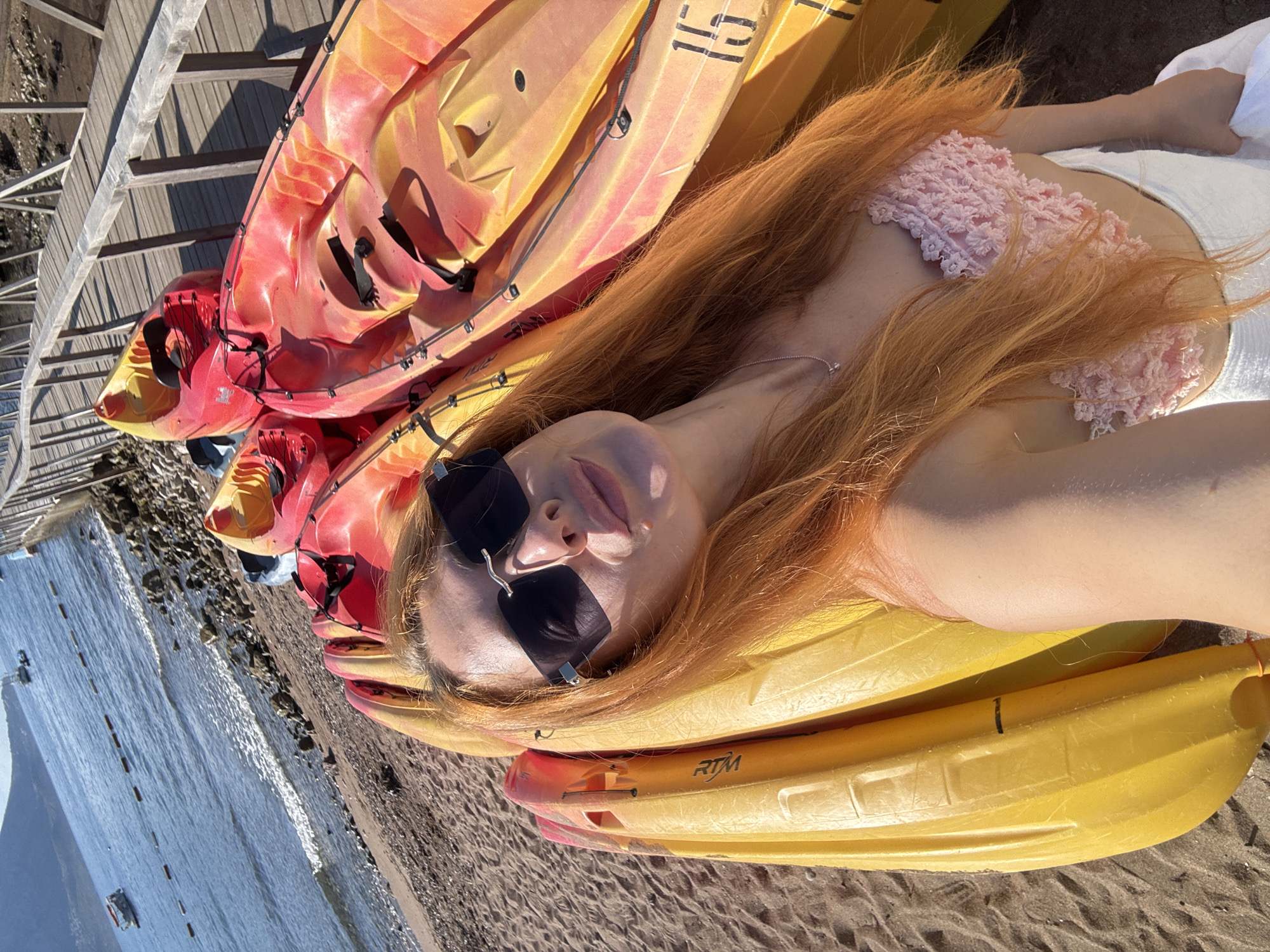 In Budva, Montenegro, a woman with long, red hair and sunglasses poses for a selfie in front of a stack of kayaks on a sandy beach. She wears a pink crocheted bikini top and white shorts, holding a white towel. The kayaks, in shades of red and yellow, are positioned on the shore, with the calm blue sea and a distant hillside visible in the background. A wooden walkway with railings runs alongside the beach, casting shadows from the sunlight. Several small boats are anchored in the water.