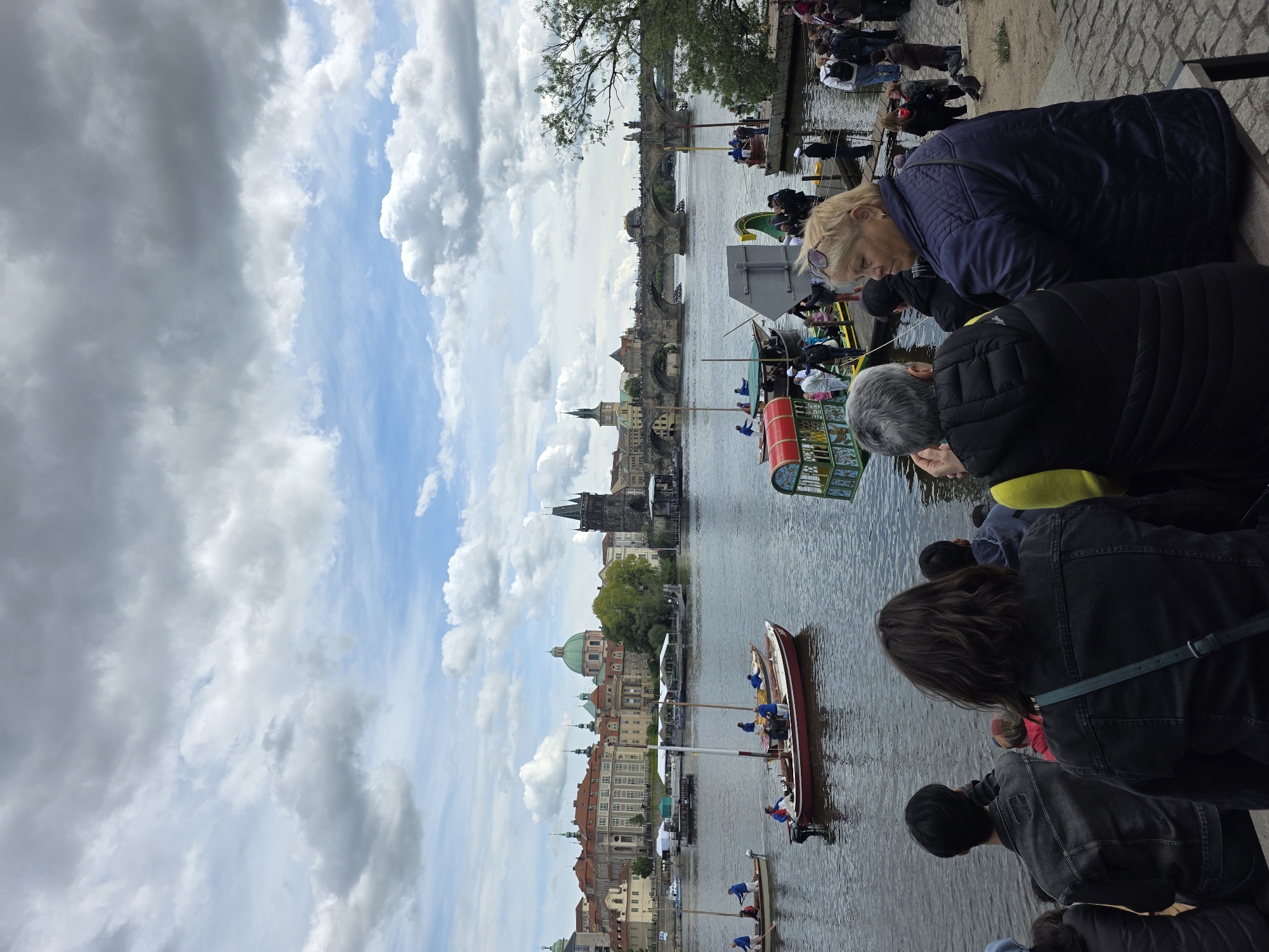The image depicts a riverside scene in Prague, Czech Republic, featuring the Vltava River and the Charles Bridge in the background. People are standing along the embankment, observing events on the water. Several narrow boats, possibly gondolas or similar vessels, are visible, with people in them. The architecture of Prague, including towers and buildings with red-tiled roofs, is visible along the riverbank. The sky is cloudy with patches of blue.