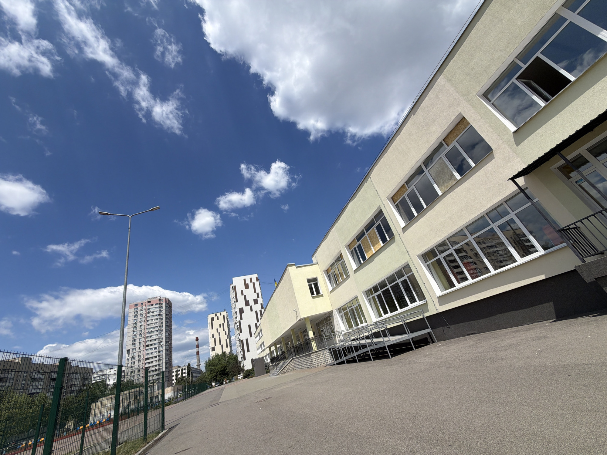 The media file depicts an exterior view of a school building with a light yellow facade, characterized by numerous large windows. The building is situated on a paved area, with a ramp leading to its entrance. In the background, several residential buildings and a tall industrial chimney are visible. The sky is a bright blue, with scattered white clouds, suggesting it is daytime. The overall scene appears to be a sunny day, possibly in the spring or summer. The perspective is low-angle, looking up towards the building and the sky. No people are visible in the media file. The location is indicated to be Kyiv, Ukraine. The image conveys a sense of an ordinary day in an urban environment.