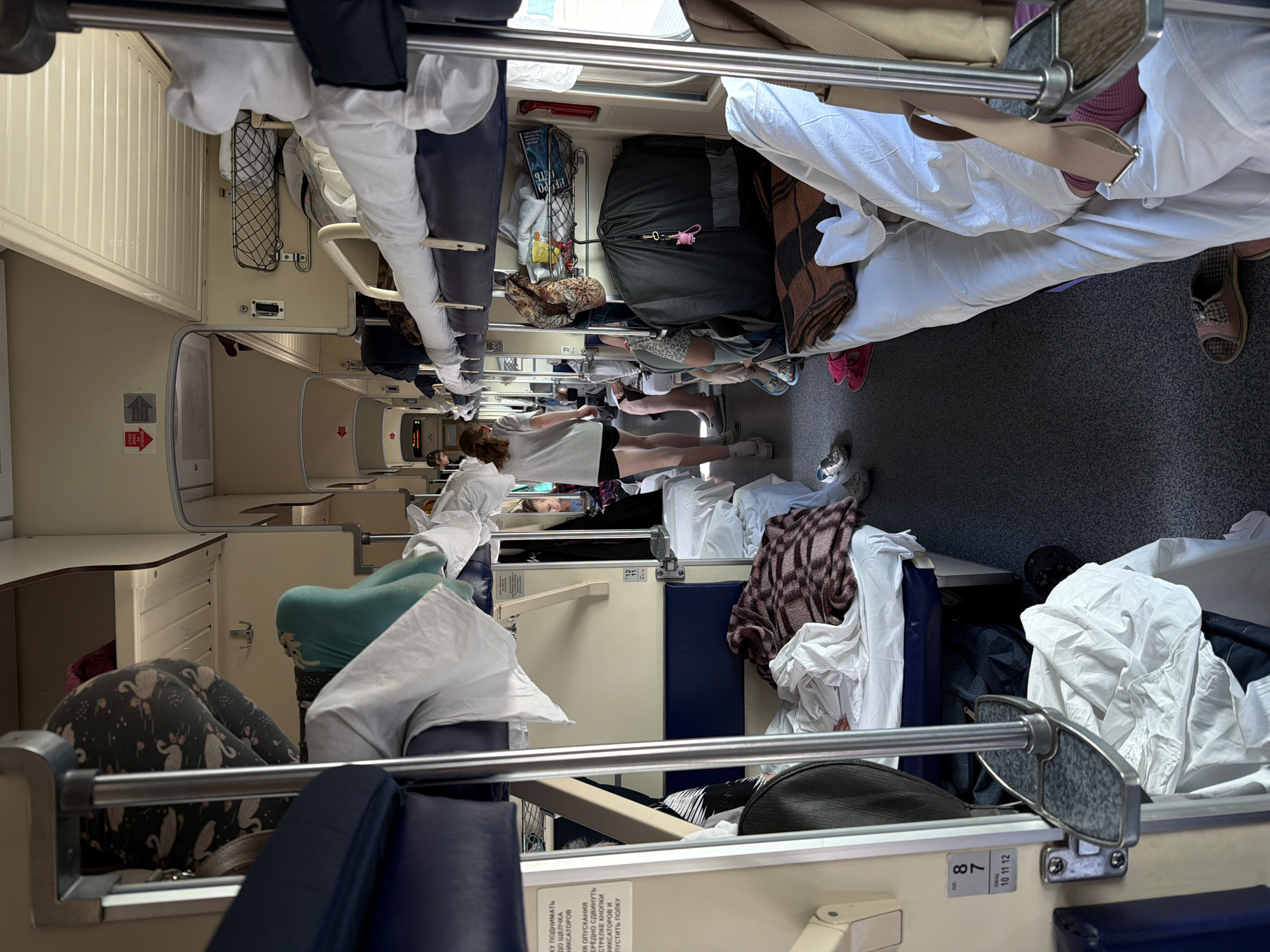 The image depicts the interior of a railway car, possibly in or around Petrov Val, Russia given Cyrillic text visible on signage. The scene appears cramped with luggage, bedding, and personal belongings filling the available space. Several people are present, including at least one person standing in the aisle, with others appearing to be seated or lying down on bunks. The railway car has multiple tiers of bunks. Overall, the scene suggests a busy and occupied railway car, possibly during a long-distance journey.