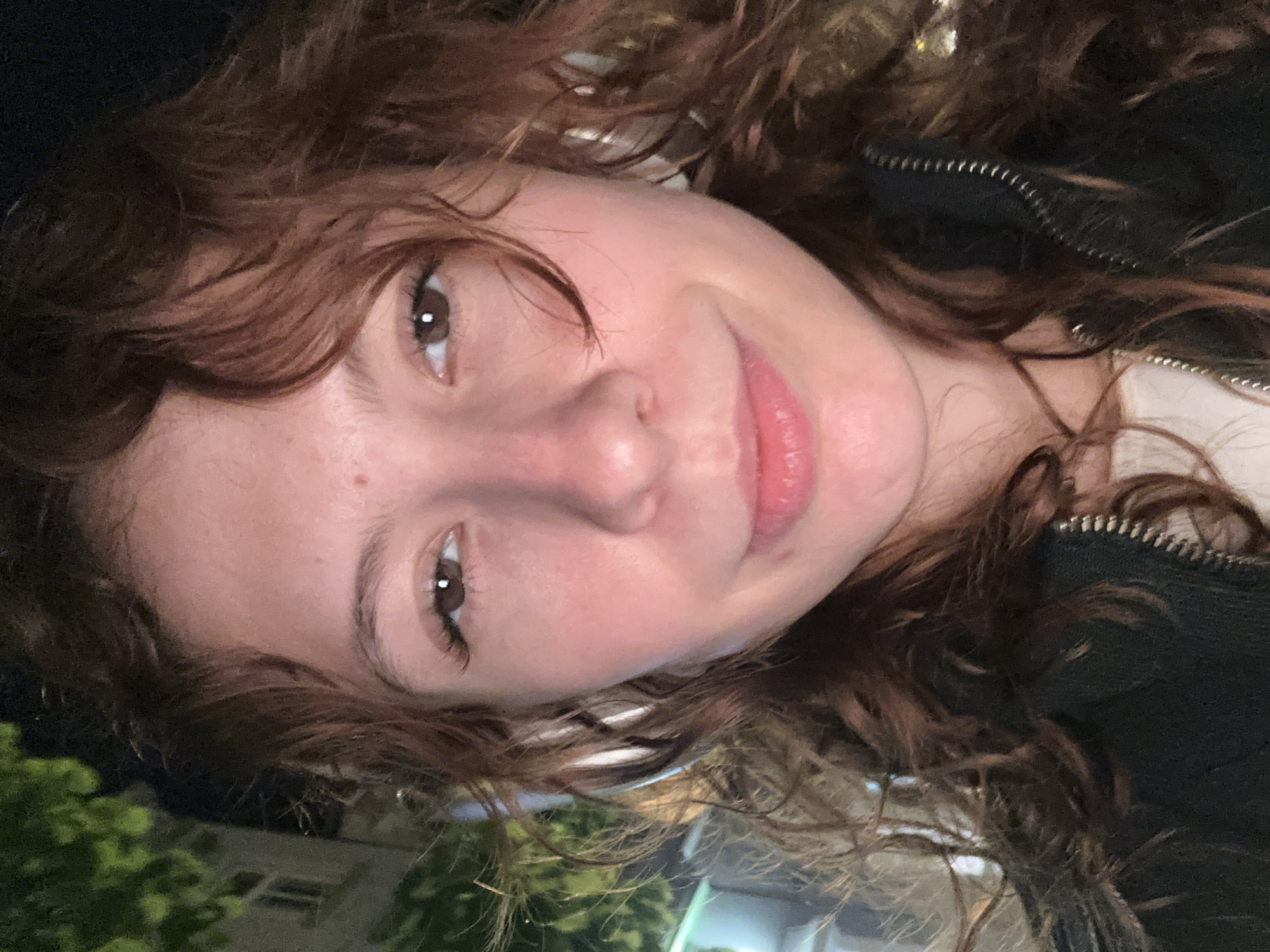 A close-up portrait features a young individual, likely female, captured with a rotated perspective. Her head is tilted significantly to the right, and she gazes directly at the viewer with a subtle, gentle smile. She has warm brown eyes and reddish-brown, curly or wavy hair that frames her face. Her complexion is fair, with a healthy blush on her cheeks and full, pink lips. A small freckle or mole is visible on her left cheek.
She is wearing a dark-colored jacket, possibly black, with a prominent silver zipper visible on the right side of the frame, over a lighter-toned, possibly white or cream, garment.
The background is dark, blurry, and out of focus, suggesting an outdoor setting at night. On the left side, indistinct green foliage, likely from trees or bushes, can be seen. Further in the background, faint, blurry shapes suggest urban structures or streetlights. The overall impression is a casual, relaxed moment. There is no visible text in the image. The scene is set in Novi Sad, Serbia.