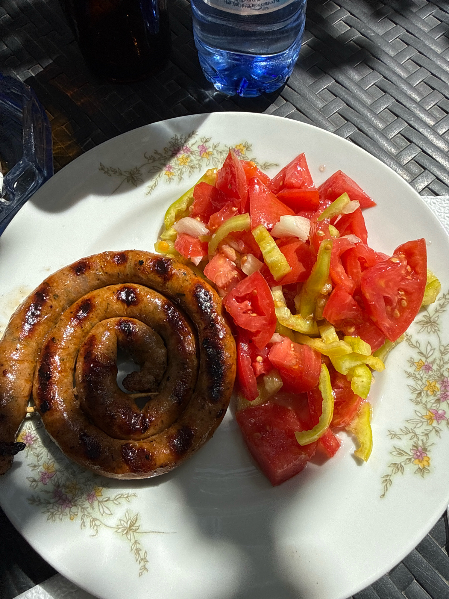 The image shows a coiled, grilled sausage and a tomato and pepper salad on a white plate with a floral pattern. The plate is on a dark, textured surface, likely a table. In the background, partially visible, are a dark glass bottle and a clear plastic bottle of water. The scene appears to be outdoors, suggested by the lighting and the textured surface which could be an outdoor table setting. The image is a close-up, focusing on the food.