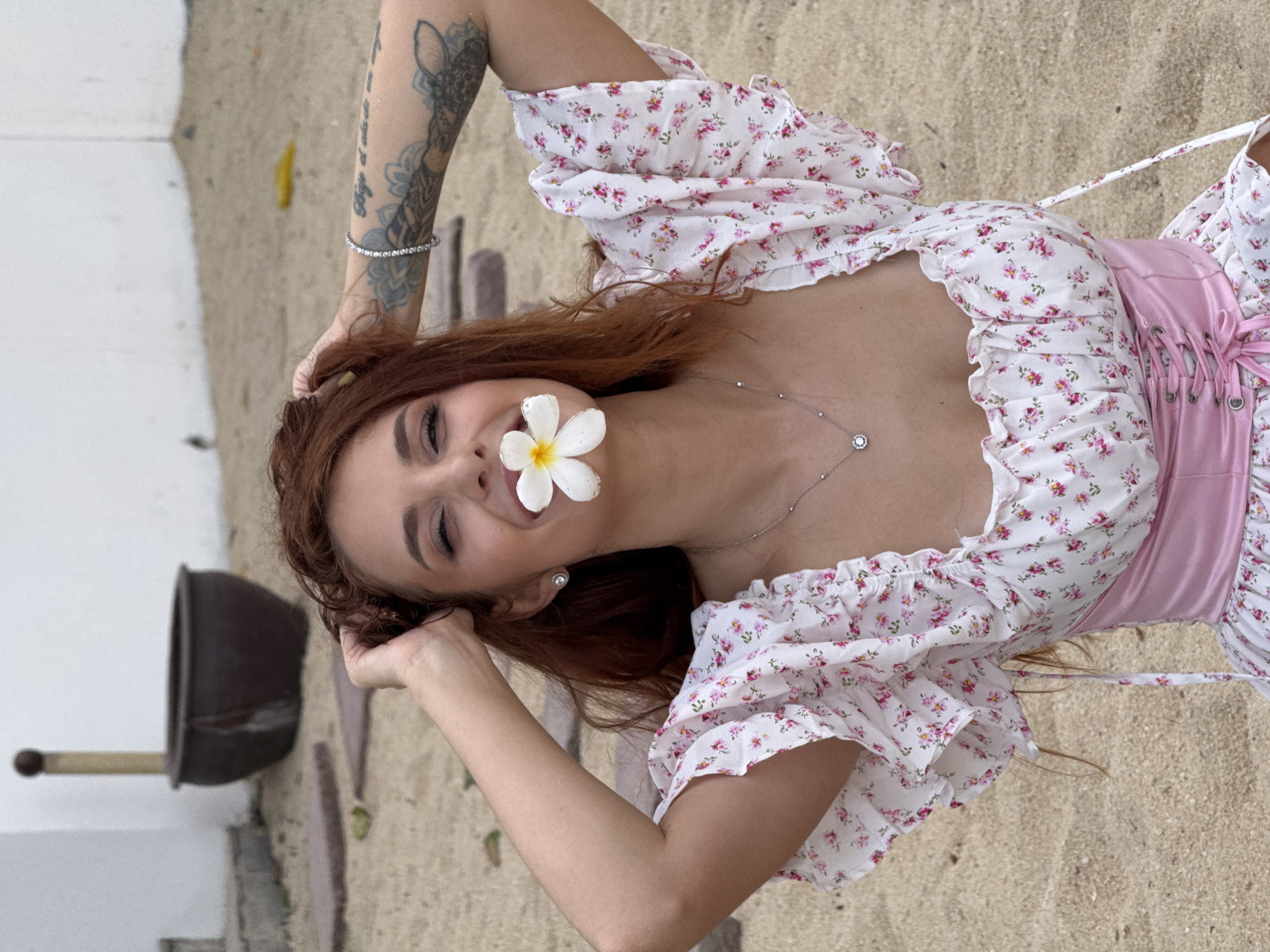 A young woman with auburn hair is outdoors on a sandy surface, possibly a beach or courtyard. She is wearing a white floral dress with pink accents and a pink corset. A white flower is held between her teeth, and her hands are clasped behind her head. A bracelet is visible on her left arm, which also displays tattoos. In the background, there is a white wall, a large dark pot, and some stone paving. The lighting suggests it is daytime. The location is identified as Ko Samui District, Thailand.