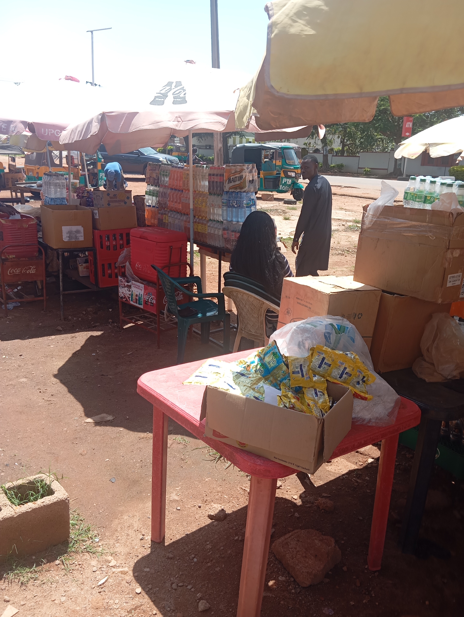 The image depicts an outdoor market stall, likely in the city of Jos, Nigeria, selling bottled drinks and packaged goods. A red plastic table in the foreground displays several boxes containing what appear to be individually packaged snack items, possibly biscuits or candies, some of which are spilling out. Behind this table, a green plastic chair is occupied by a person facing away from the camera, with long dark hair. To the left of the chair, a red cooler with the "Coca-Cola" logo is visible, alongside stacks of Coca-Cola crates.
In the mid-ground, several shelves are laden with bottled beverages, including brands like "Maltina" and other water and soft drink bottles. A man in dark clothing is standing and facing the right side of the frame, looking towards the products. Further back, a person in a blue shirt is bent over, seemingly arranging items. Various cardboard boxes are stacked on and around tables and shelves, some bearing visible labels. In the background, a three-wheeled vehicle, possibly an auto-rickshaw, is parked. The ground is a dirt surface, characteristic of outdoor markets. Overhead, umbrellas provide shade for the stalls. The overall impression is a busy, functional marketplace under a clear sky.