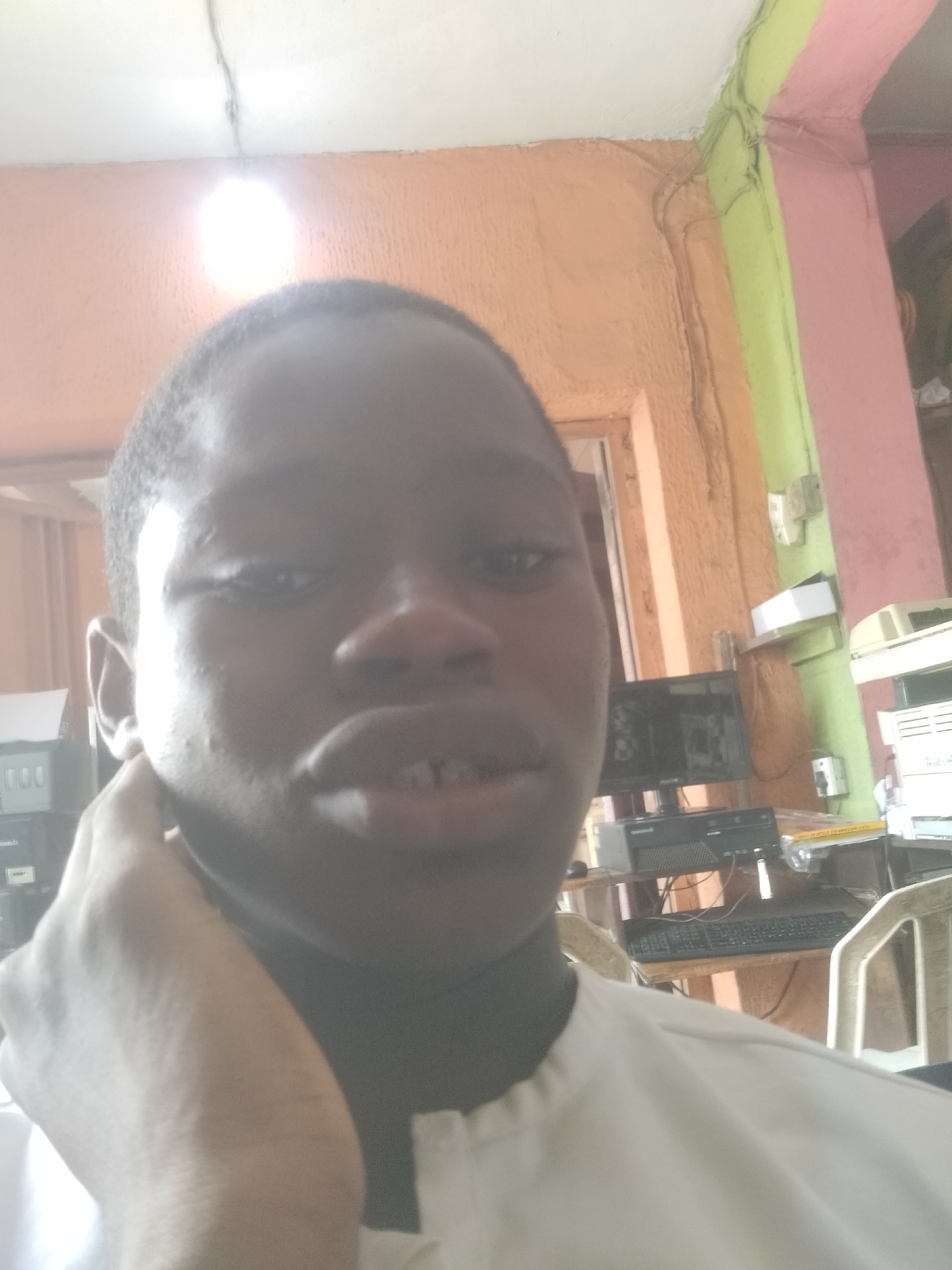 A close-up selfie of a young, dark-skinned man with closely cropped hair, possibly in his late teens or early twenties, is displayed indoors. The subject is wearing a white garment. He is touching his cheek with his left hand.
Behind the subject, the background reveals a cluttered interior scene. A desk with computer equipment, including a monitor, CPU, and keyboard, is visible. A white plastic chair sits beside the desk. The walls are painted in a mix of colors, including orange, green, and pink. The presence of wires and cables indicates possible electrical wiring.
The location is possibly Shangisha, Nigeria. No further location data is provided.